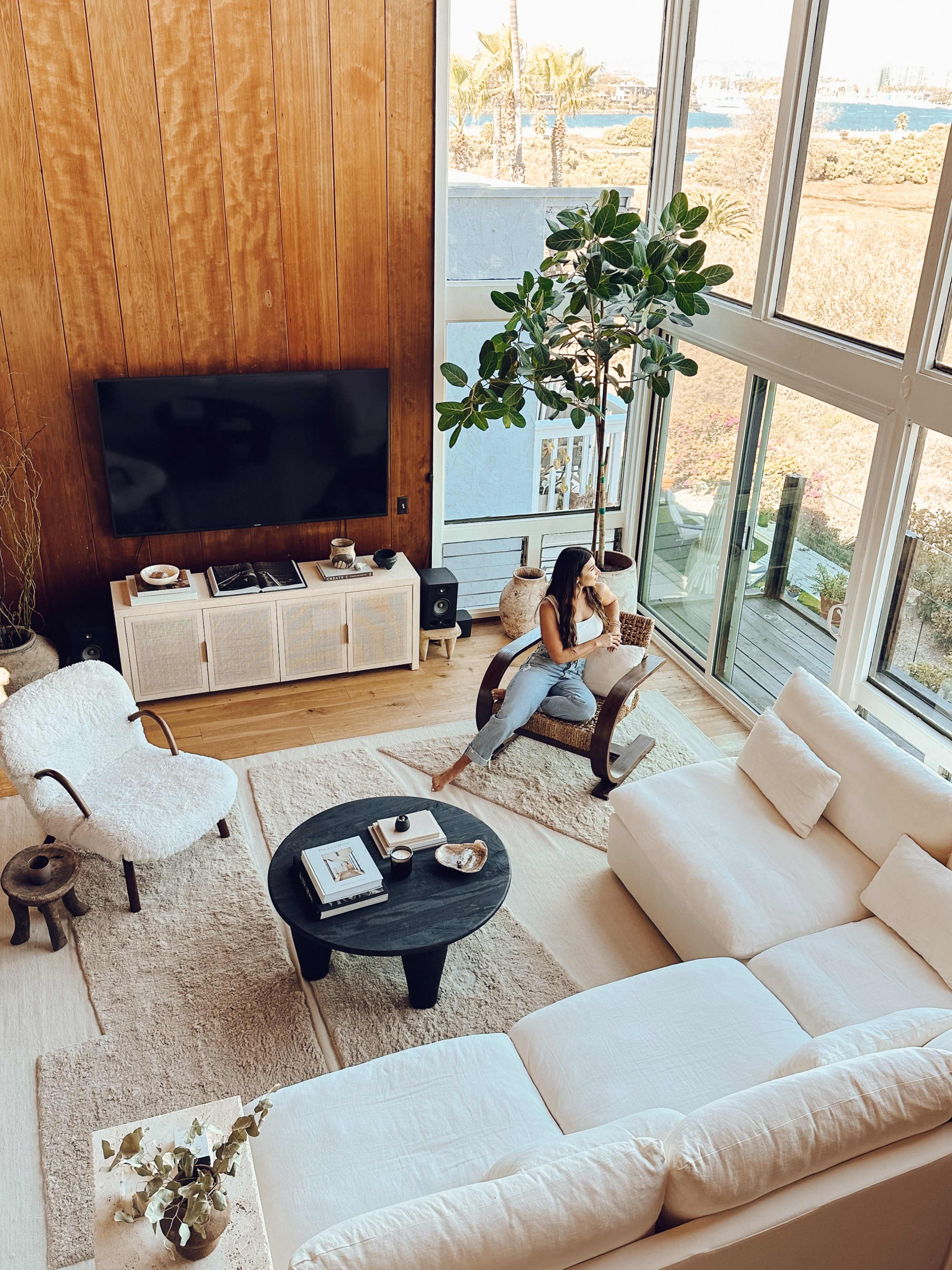 A bright living room features large windows with a view of the outdoors, a sectional sofa, a round coffee table, and a woman sitting comfortably while reading.