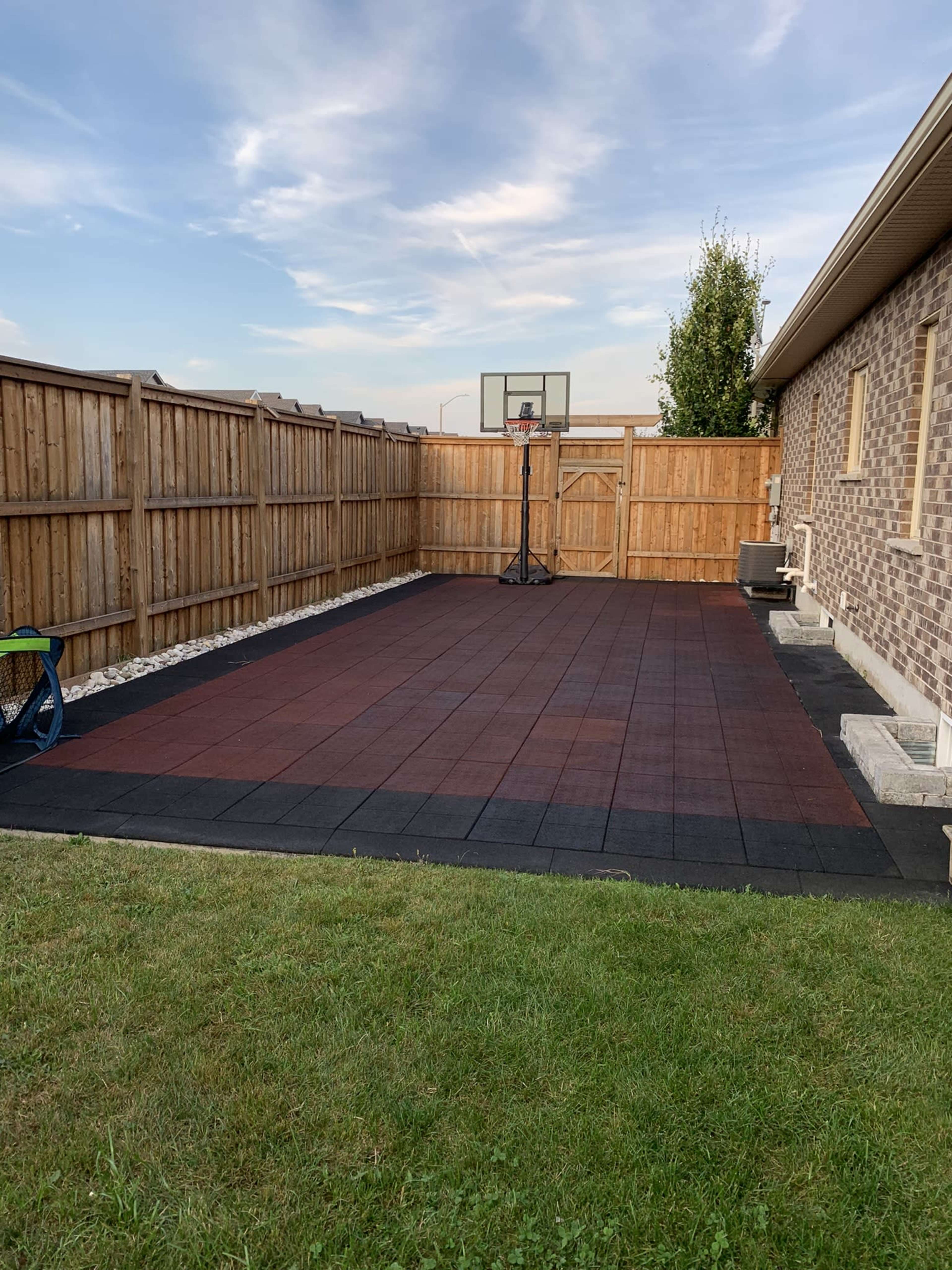 A fenced backyard with a basketball hoop mounted on a concrete slab surrounded by rubber flooring.
