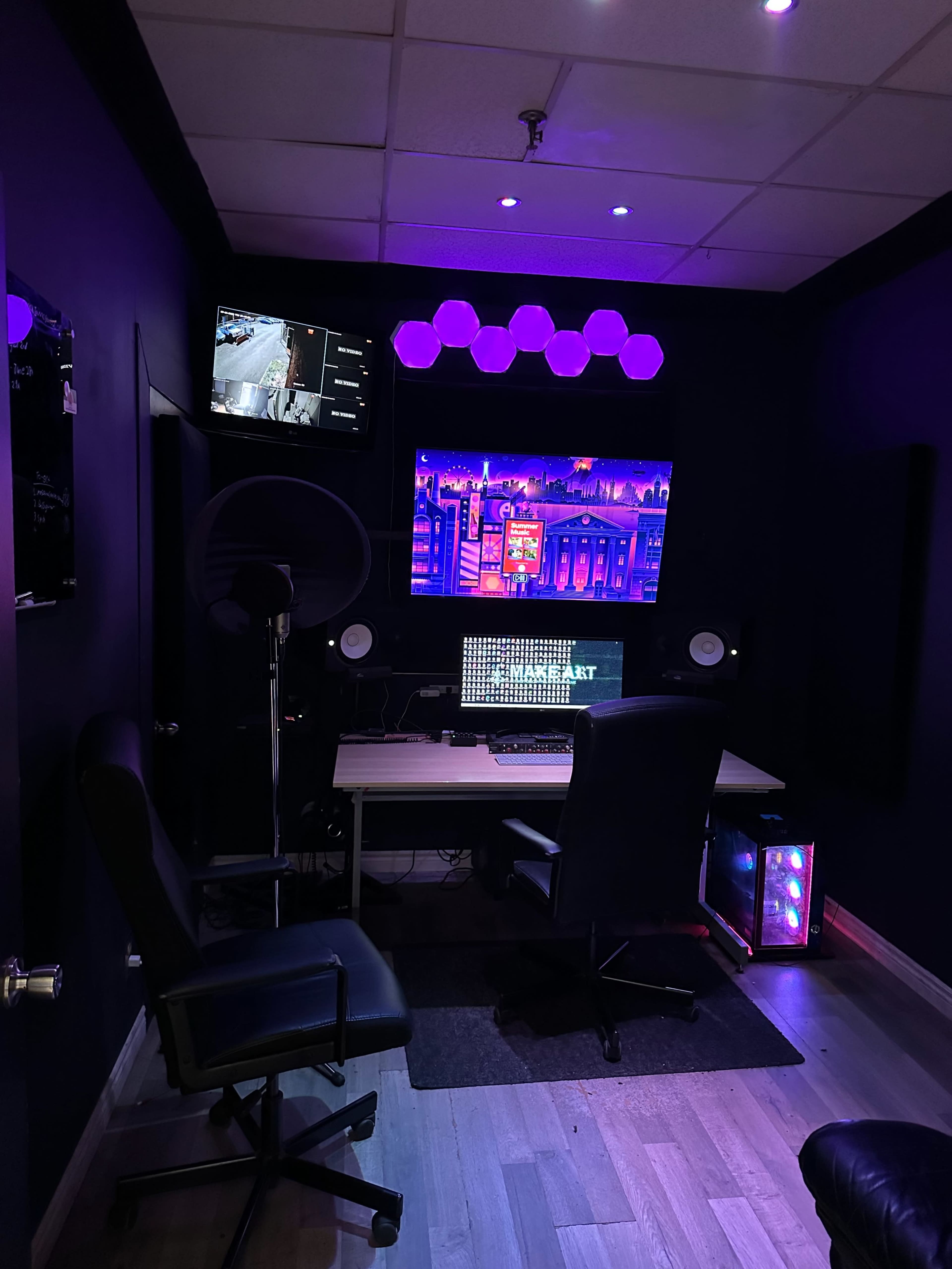 The image shows a dark room with a desk containing a computer setup, a monitor displaying a colorful screen, and ambient purple lighting.