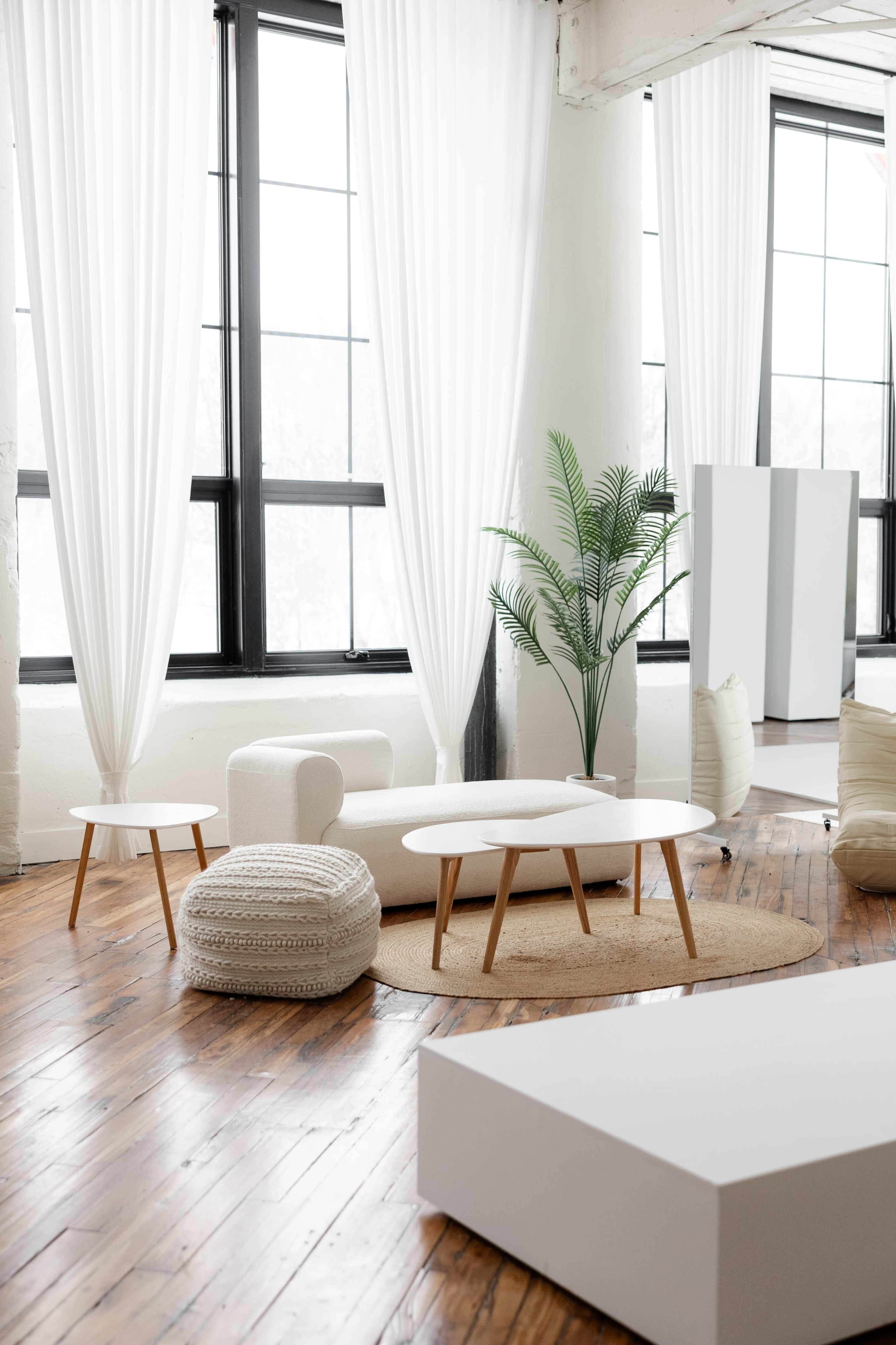 The image shows a modern, minimalistic living space with a white sofa, a round coffee table, and a plant, all arranged on a wooden floor with large windows and sheer curtains.