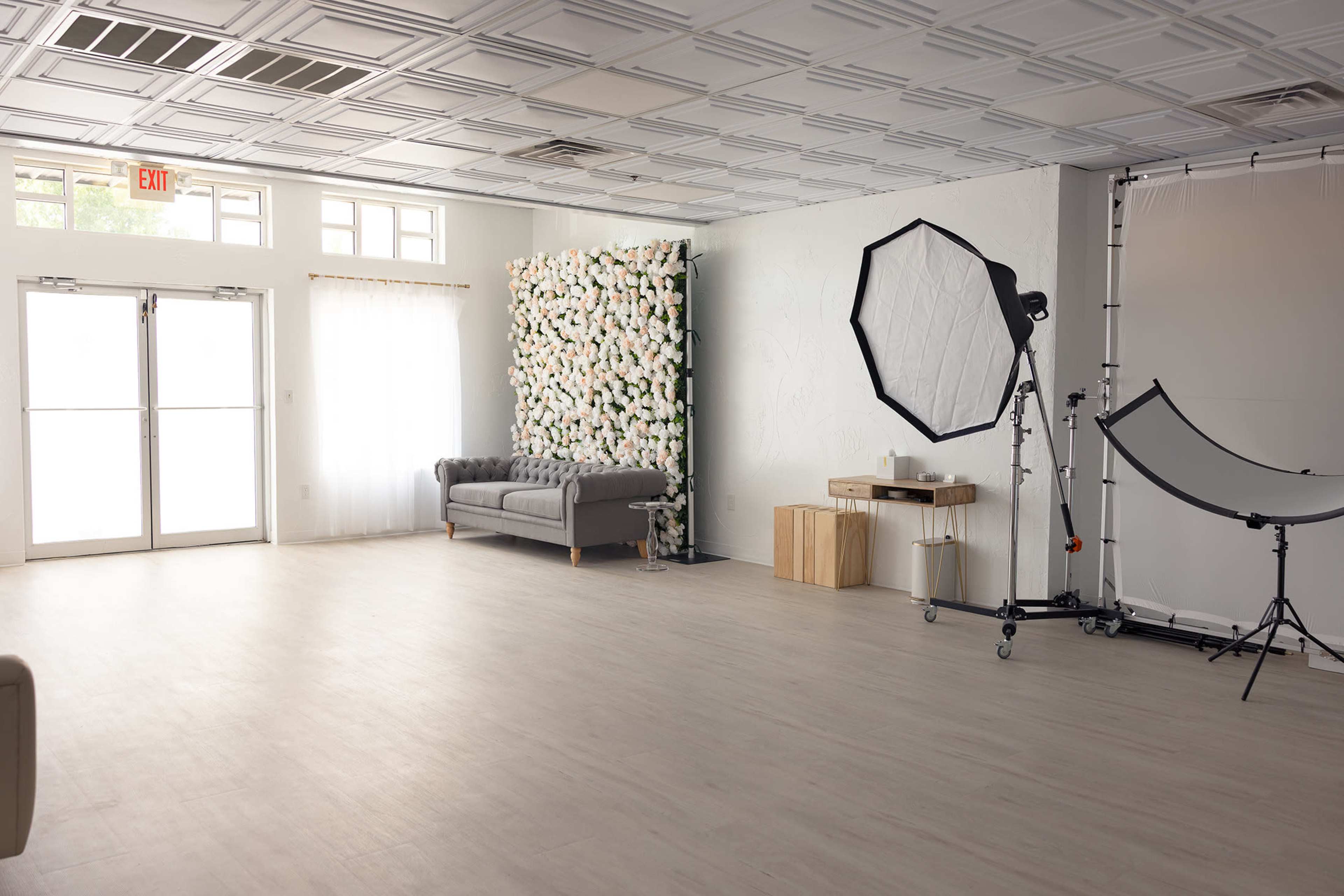 The image shows a spacious interior with a light-colored floor, a floral wall decoration, a gray sofa, and photography equipment set up in a corner.