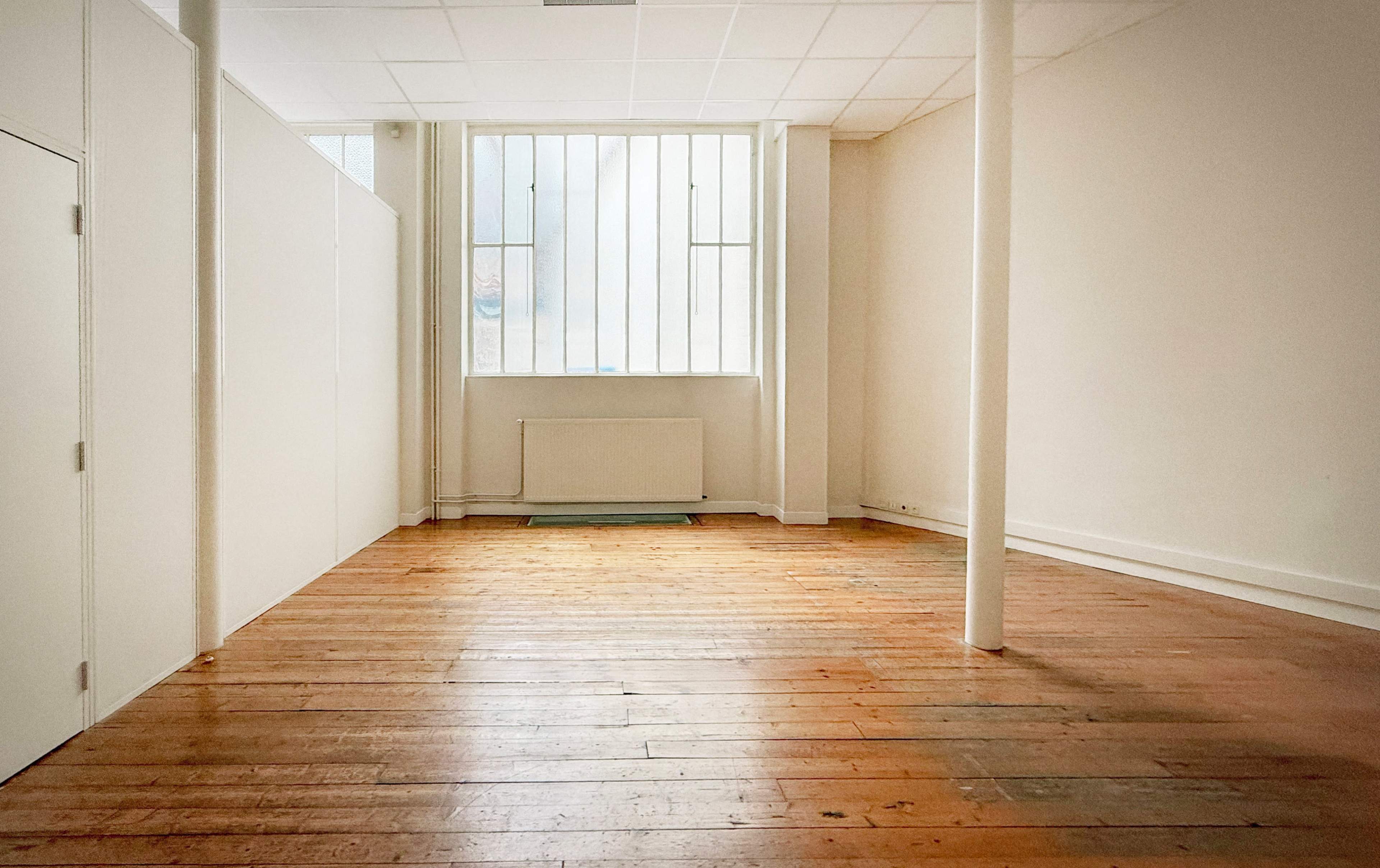 The image shows a spacious, empty room with wooden floors, a large window, and white walls.