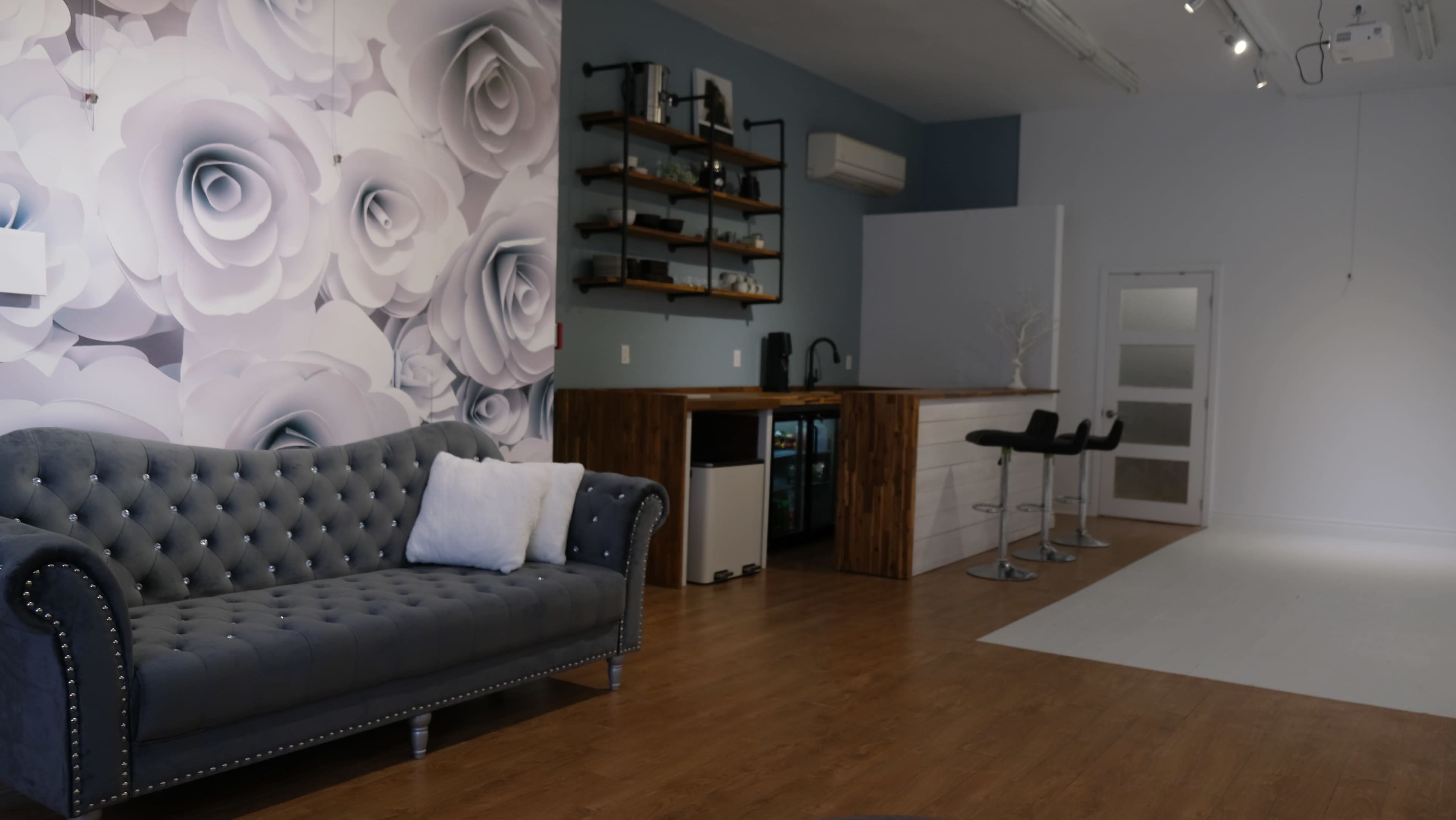A modern interior featuring a gray tufted sofa, a floral wall mural, a wooden bar area with stools, and a small kitchen setup in a spacious room with hardwood flooring.