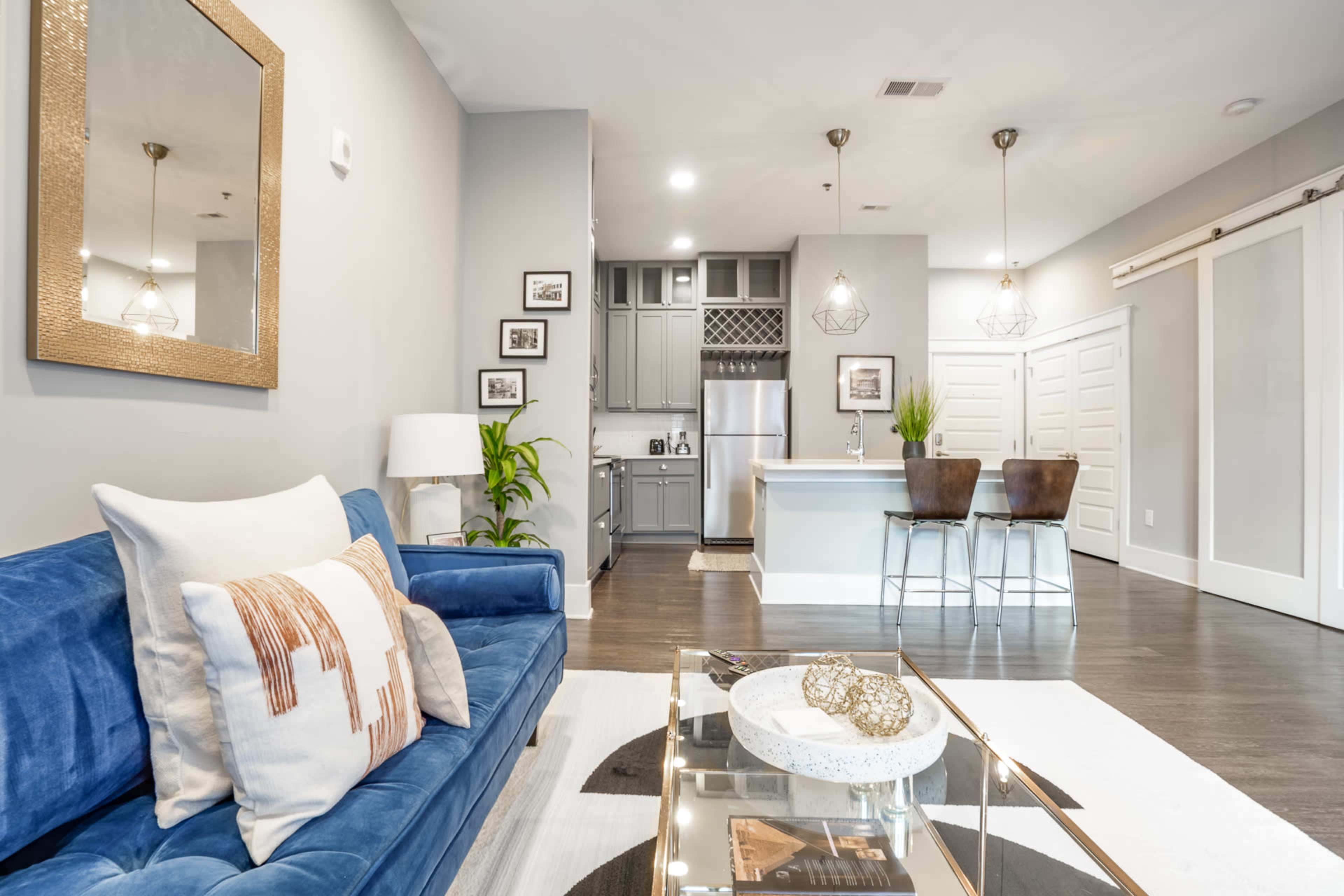 The image shows a modern living room featuring a blue sofa, a glass coffee table, and an open kitchen area with gray cabinetry and bar stools.