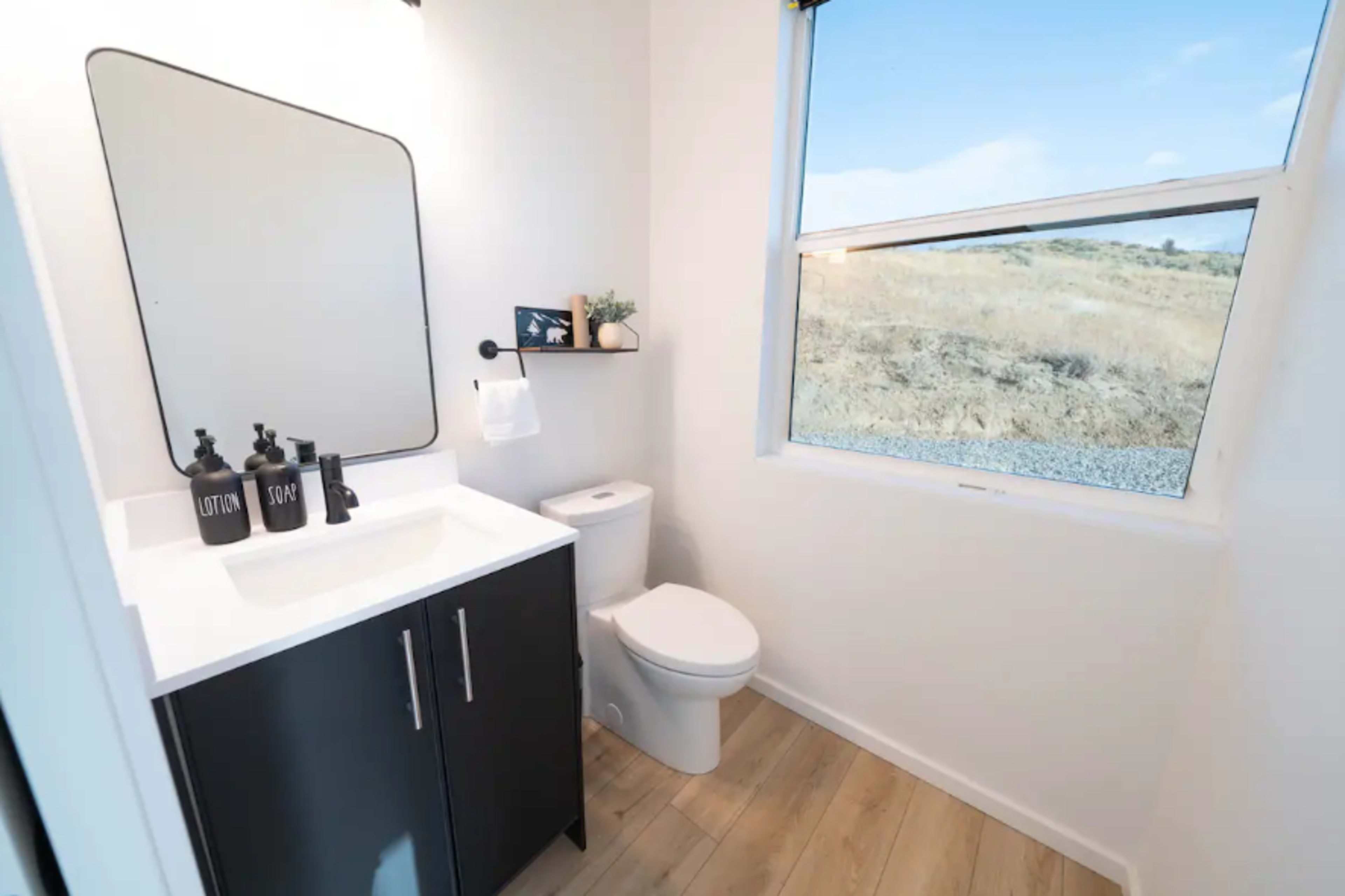 A modern bathroom with a white sink and black cabinet, a toilet, and a window revealing a view of the dry landscape outside.