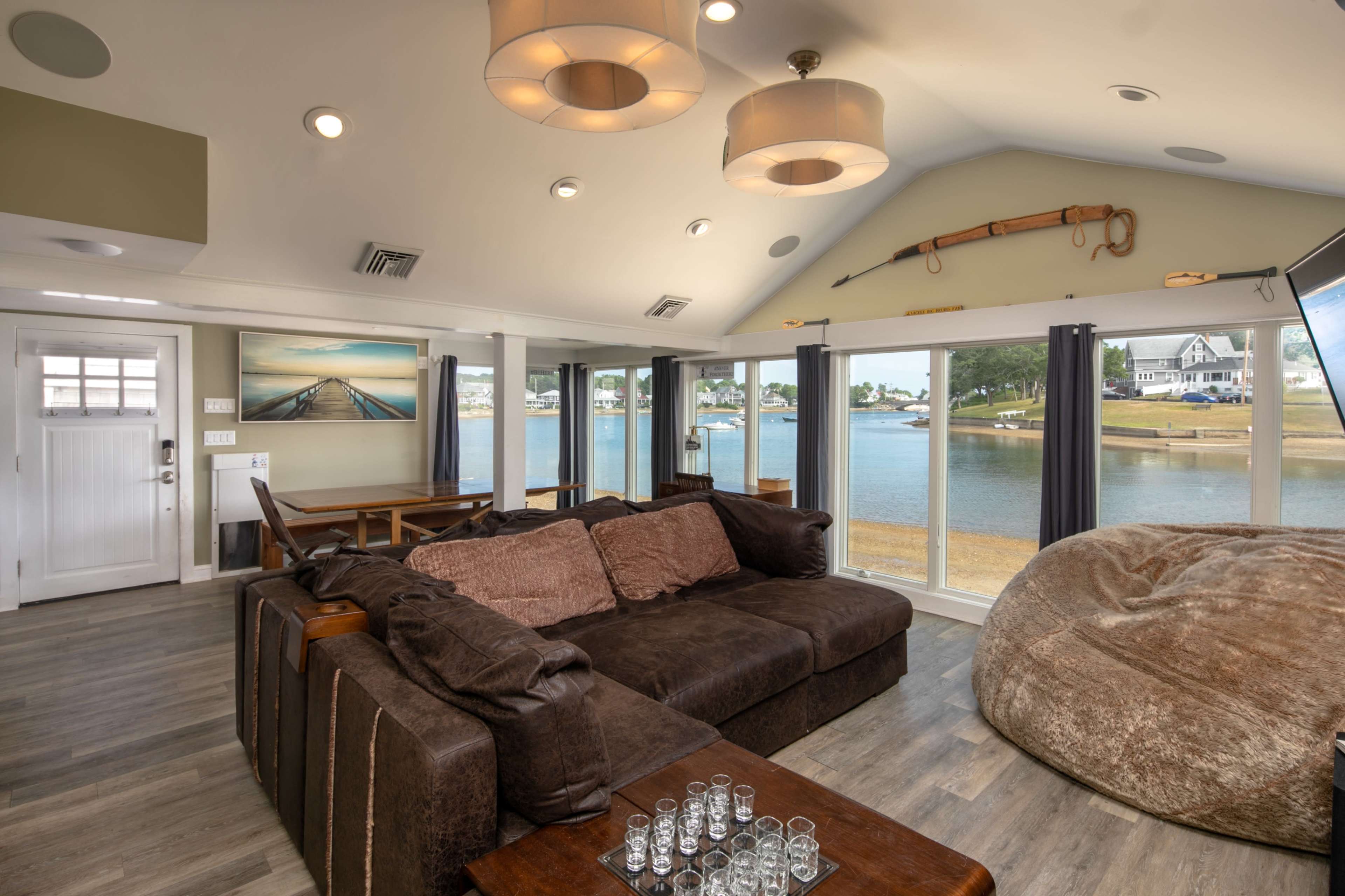 The interior of a modern living room features a brown sectional sofa, a dining area, large windows overlooking a water view, and various decorative items on the walls.