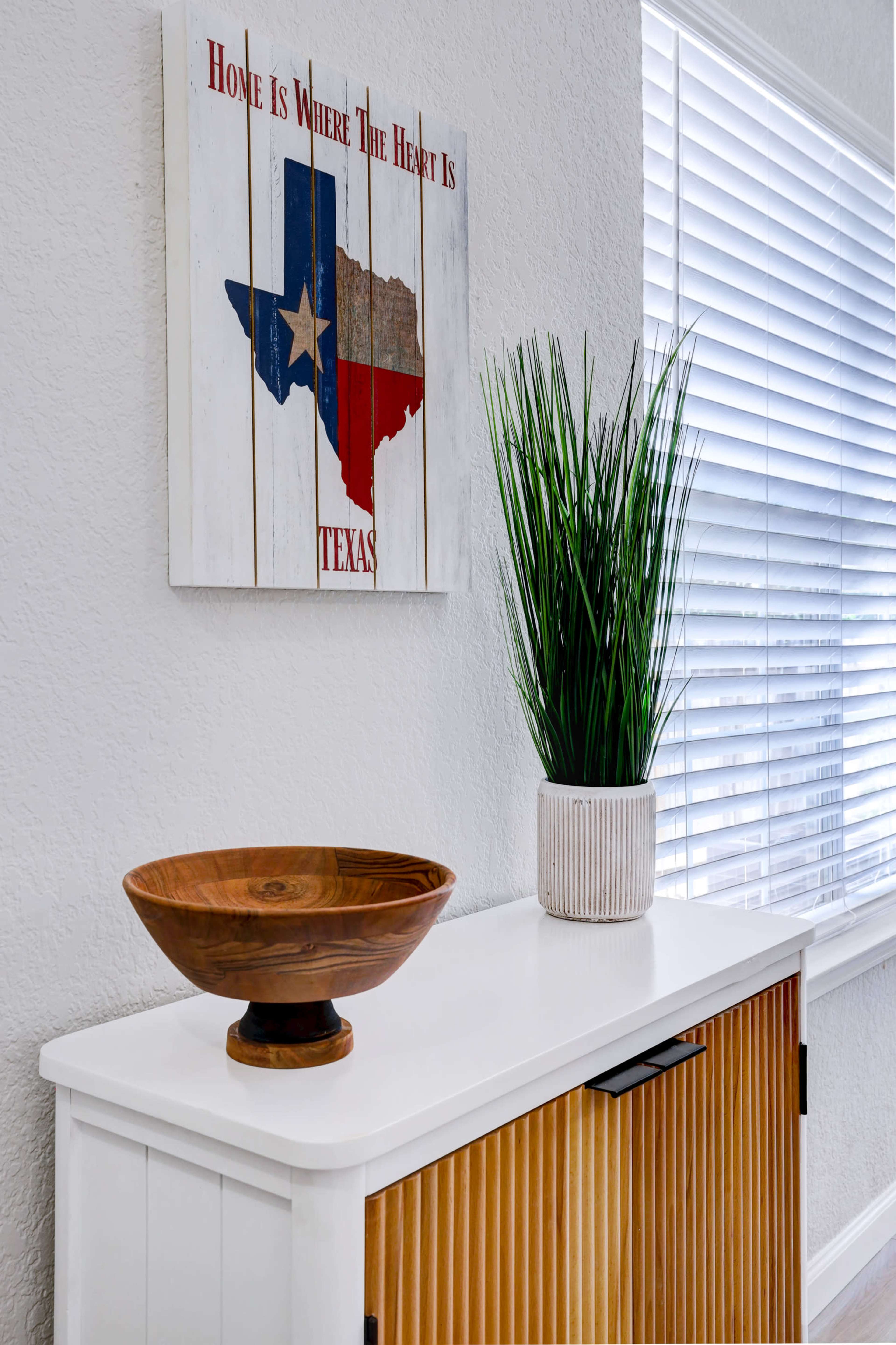 A wooden bowl sits on a white dresser beneath a wall art piece depicting the Texas flag, next to a potted plant, with blinds filtering sunlight from a window.