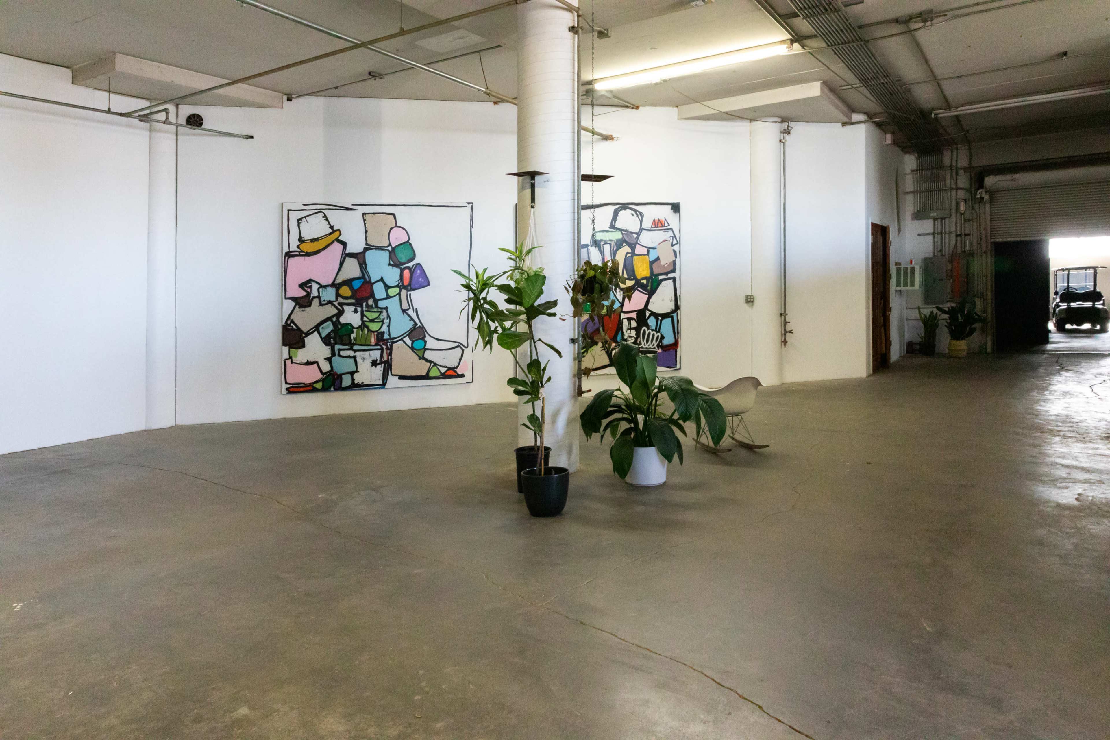 An expansive, open interior space with concrete flooring, two abstract paintings on the wall, and several potted plants.