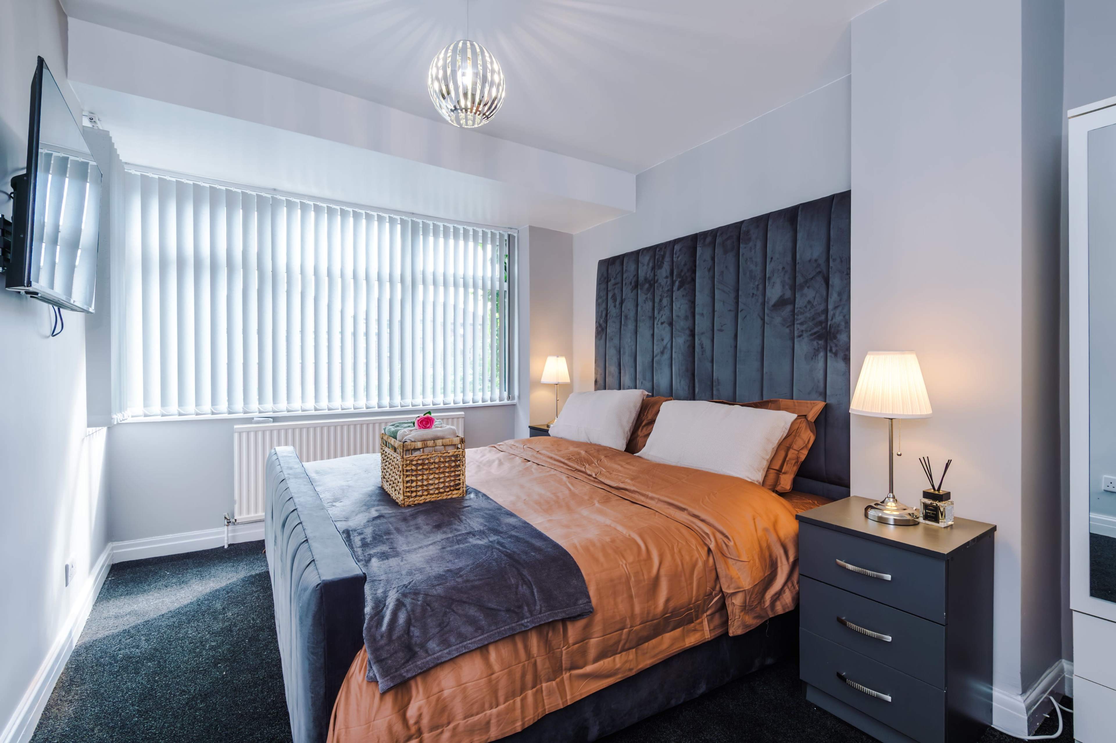 A well-furnished bedroom features a large bed with an orange and gray duvet, a bedside table with a lamp, a wall-mounted TV, and vertical blinds allowing light to enter from the window.
