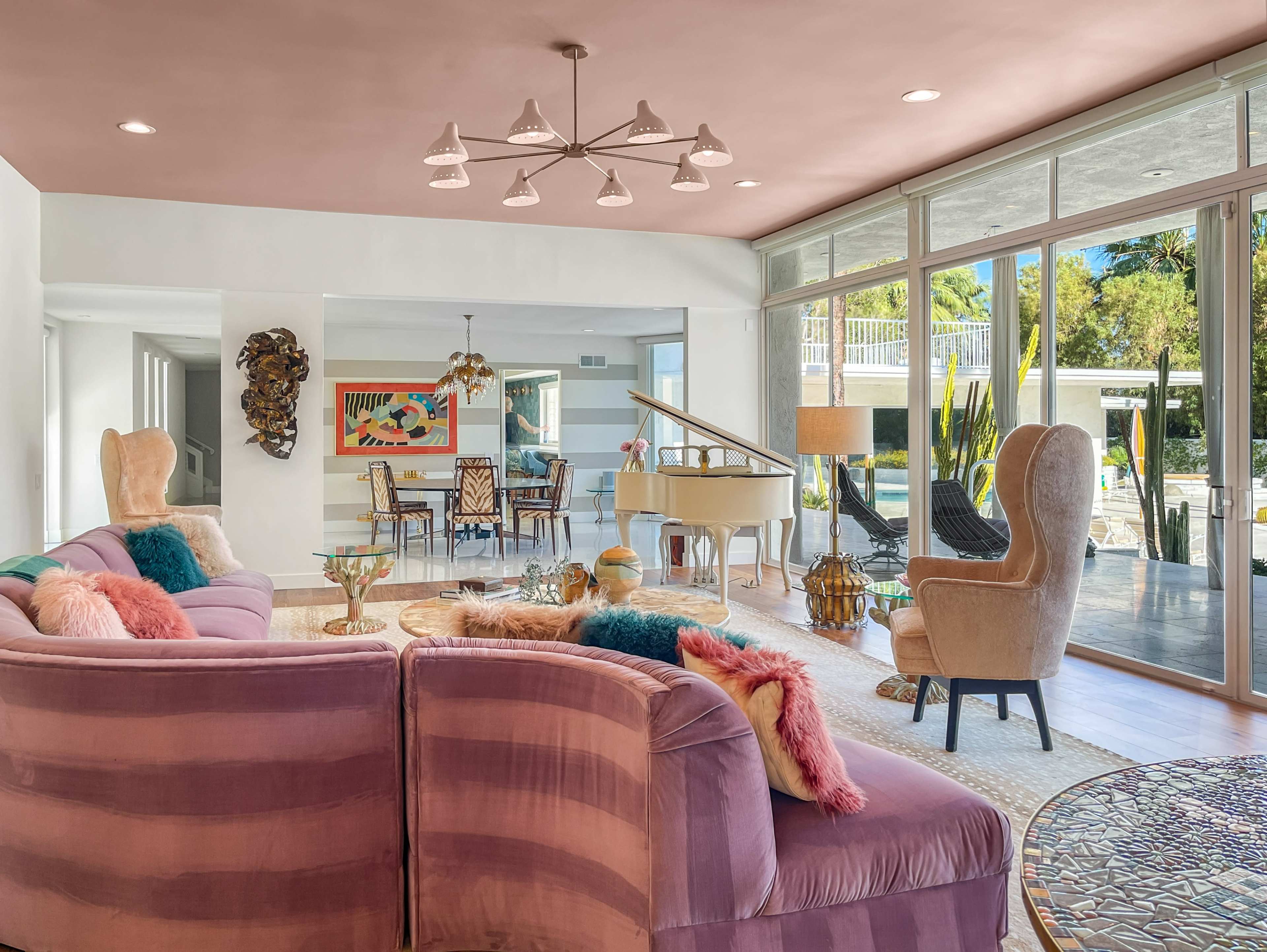 A modern living room featuring a grand piano, a pink sofa, and large windows overlooking an outdoor area.