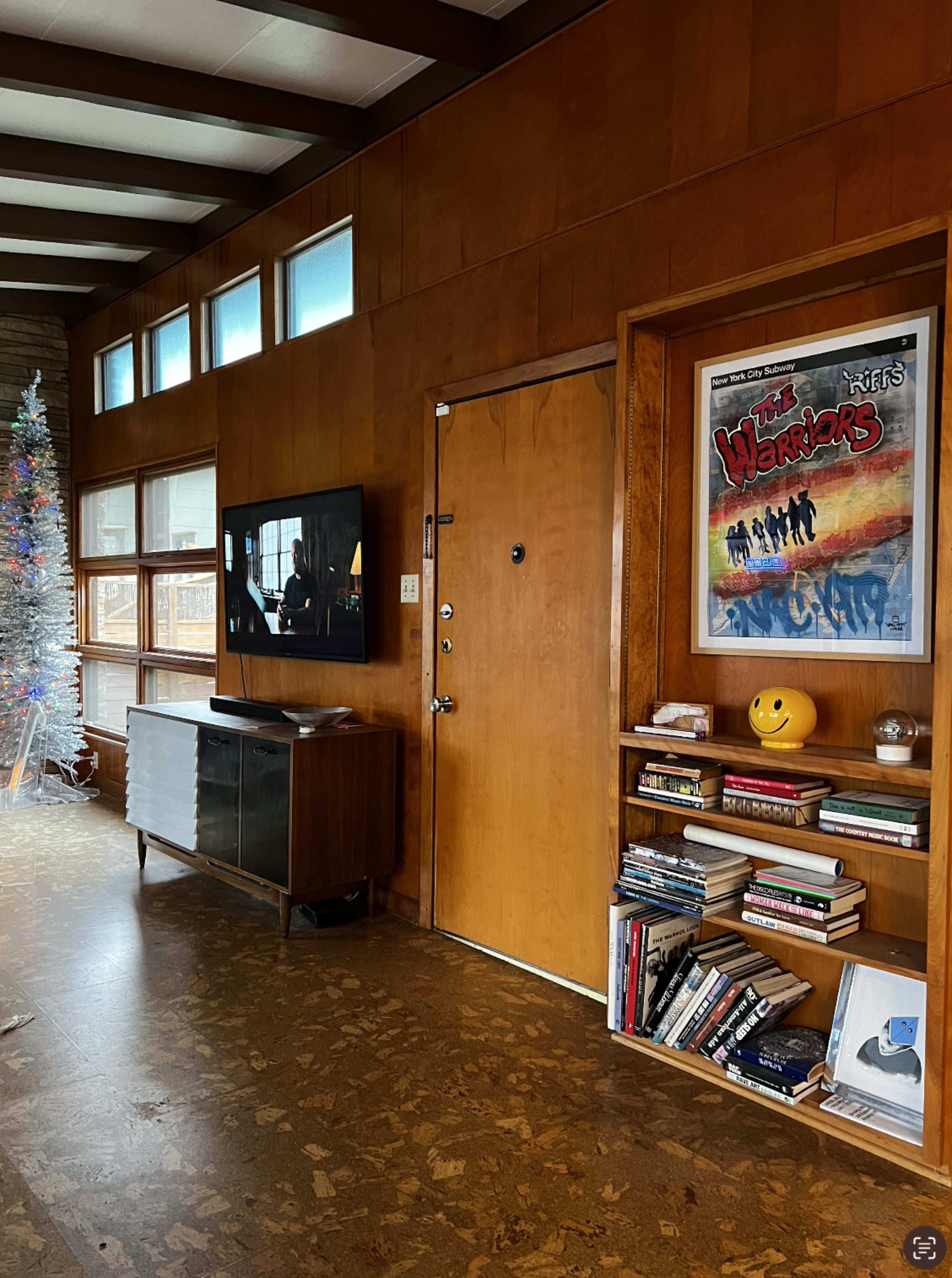The image shows a living room with wooden paneling, a television mounted on the wall, a door with a small window, a bookshelf filled with books and a smiling yellow toy, and a Christmas tree in the corner.