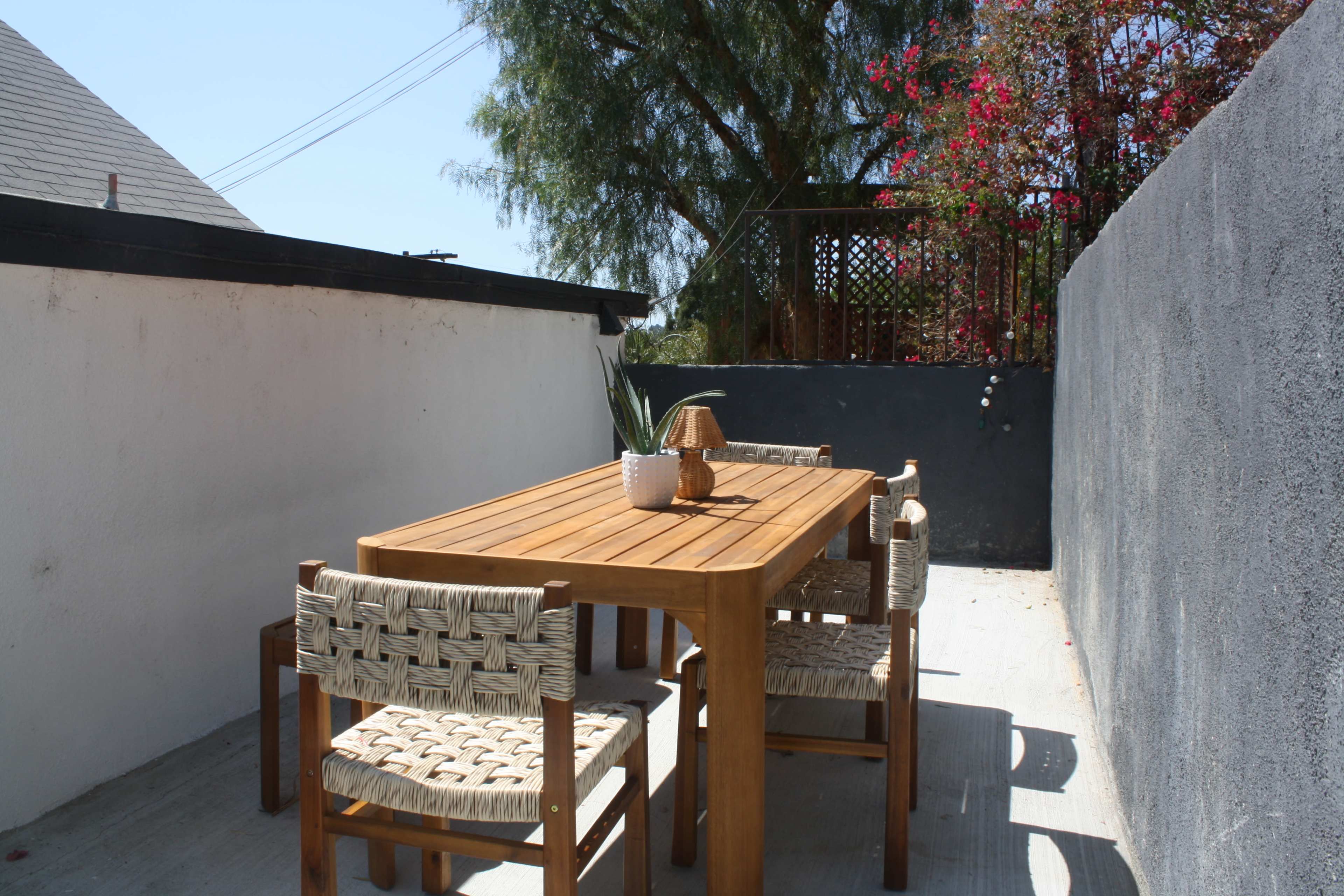 A rectangular wooden dining table with four chairs is set in a bright outdoor space surrounded by a wall, with flowering plants in the background.