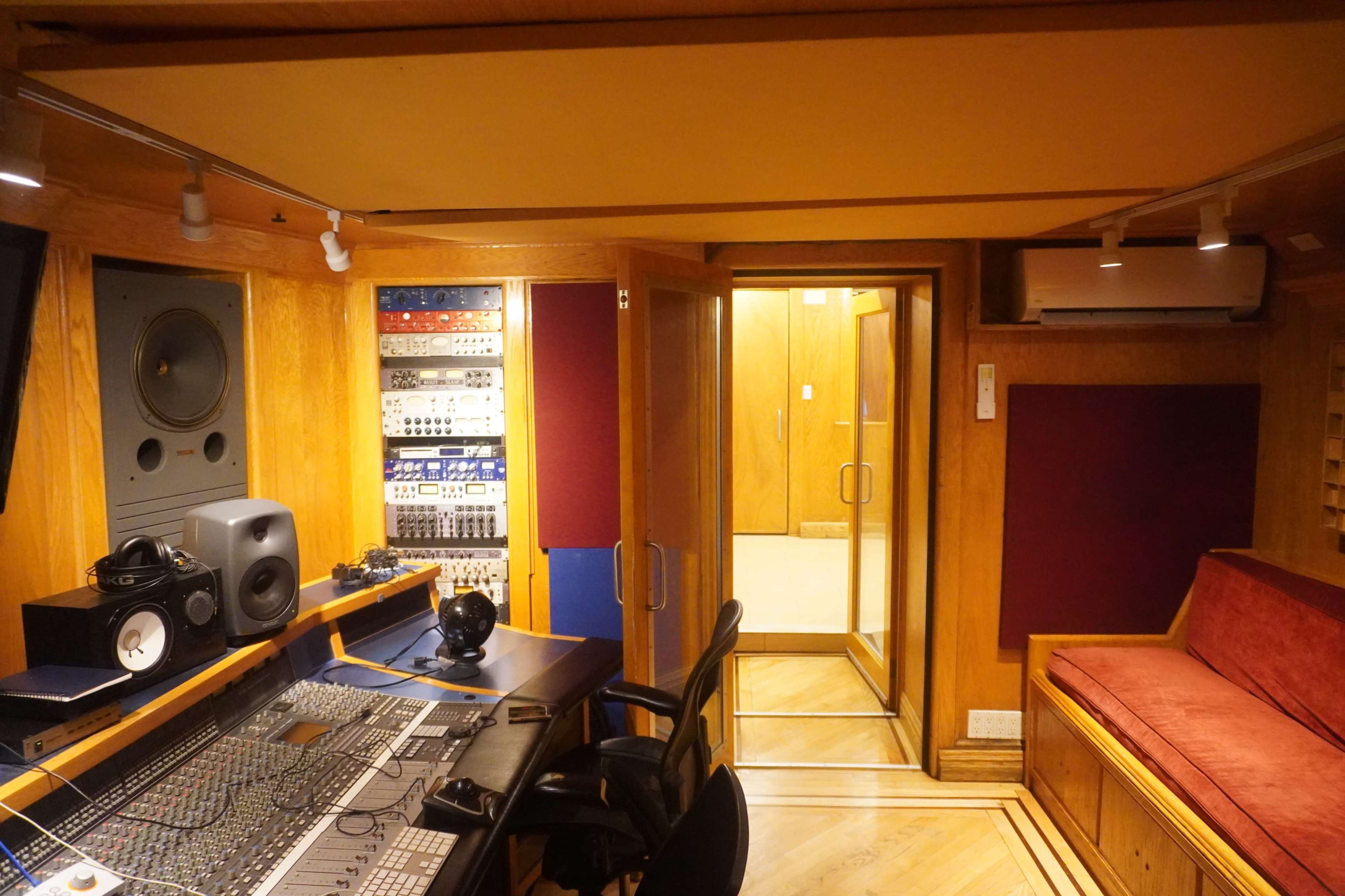 A sound studio featuring a control desk with audio equipment, a red couch, and a glass door leading to an adjacent room.