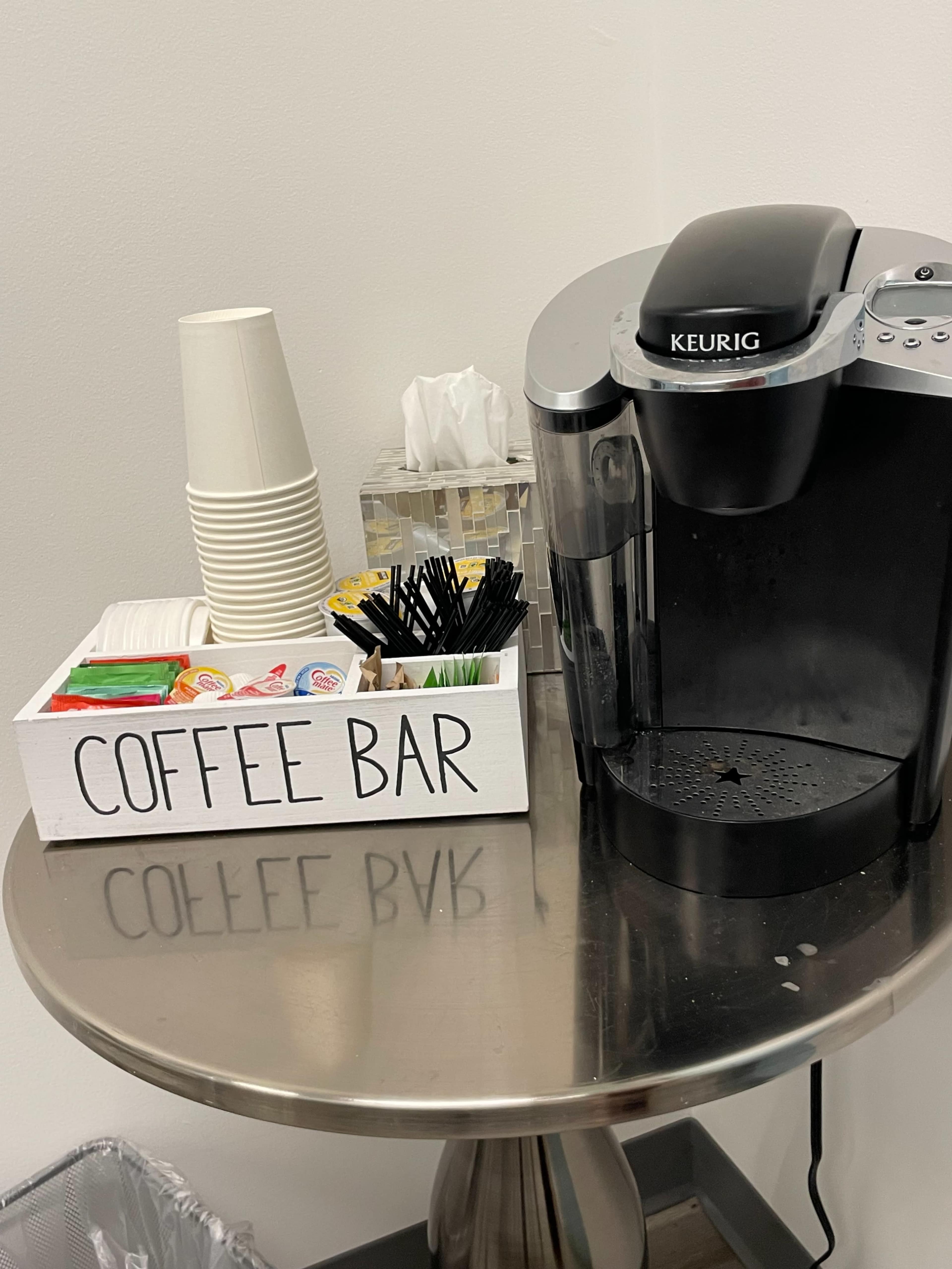 A coffee bar setup featuring a Keurig machine, disposable cups, and a tray with assorted coffee condiments and stirrers.