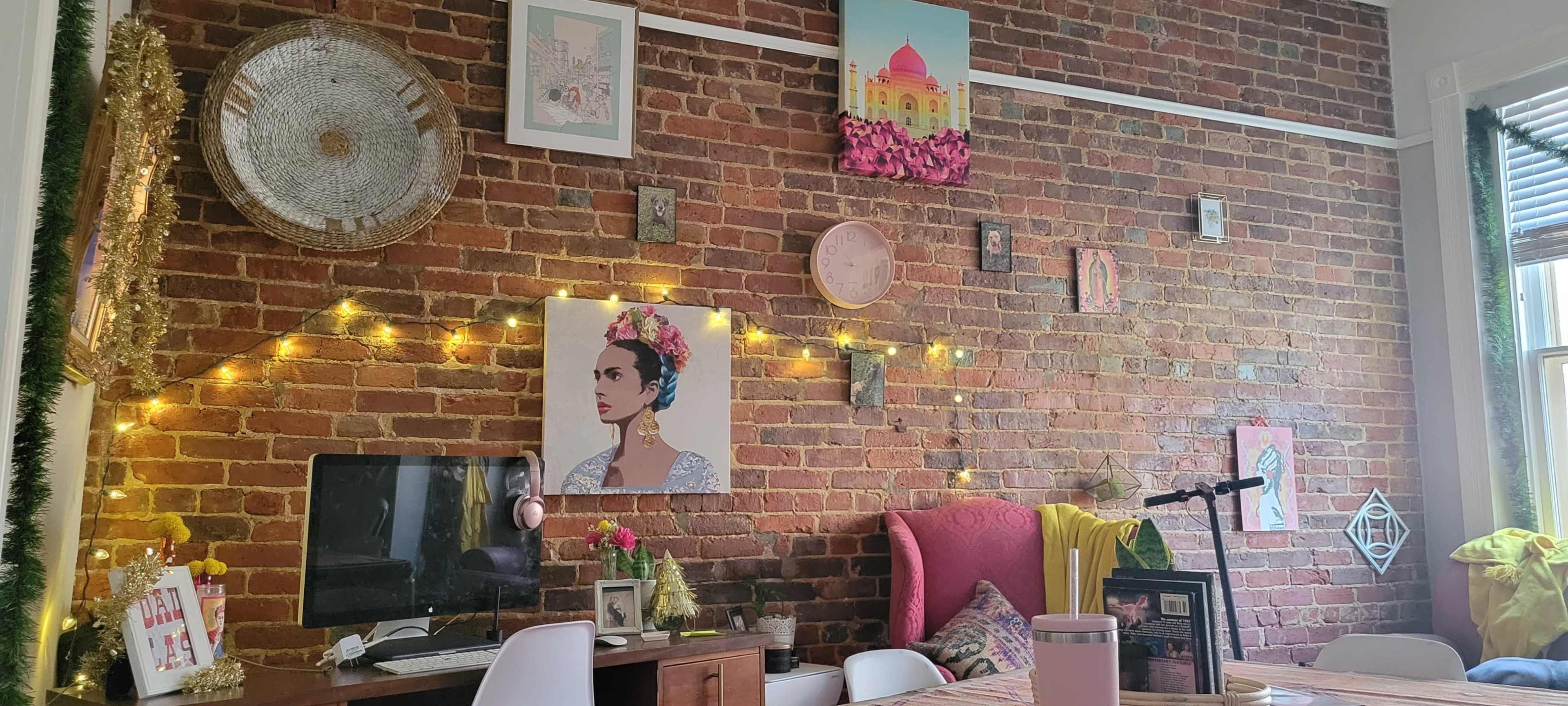 A brick wall adorned with various framed artworks, fairy lights, and a workspace featuring a computer and a pink chair.