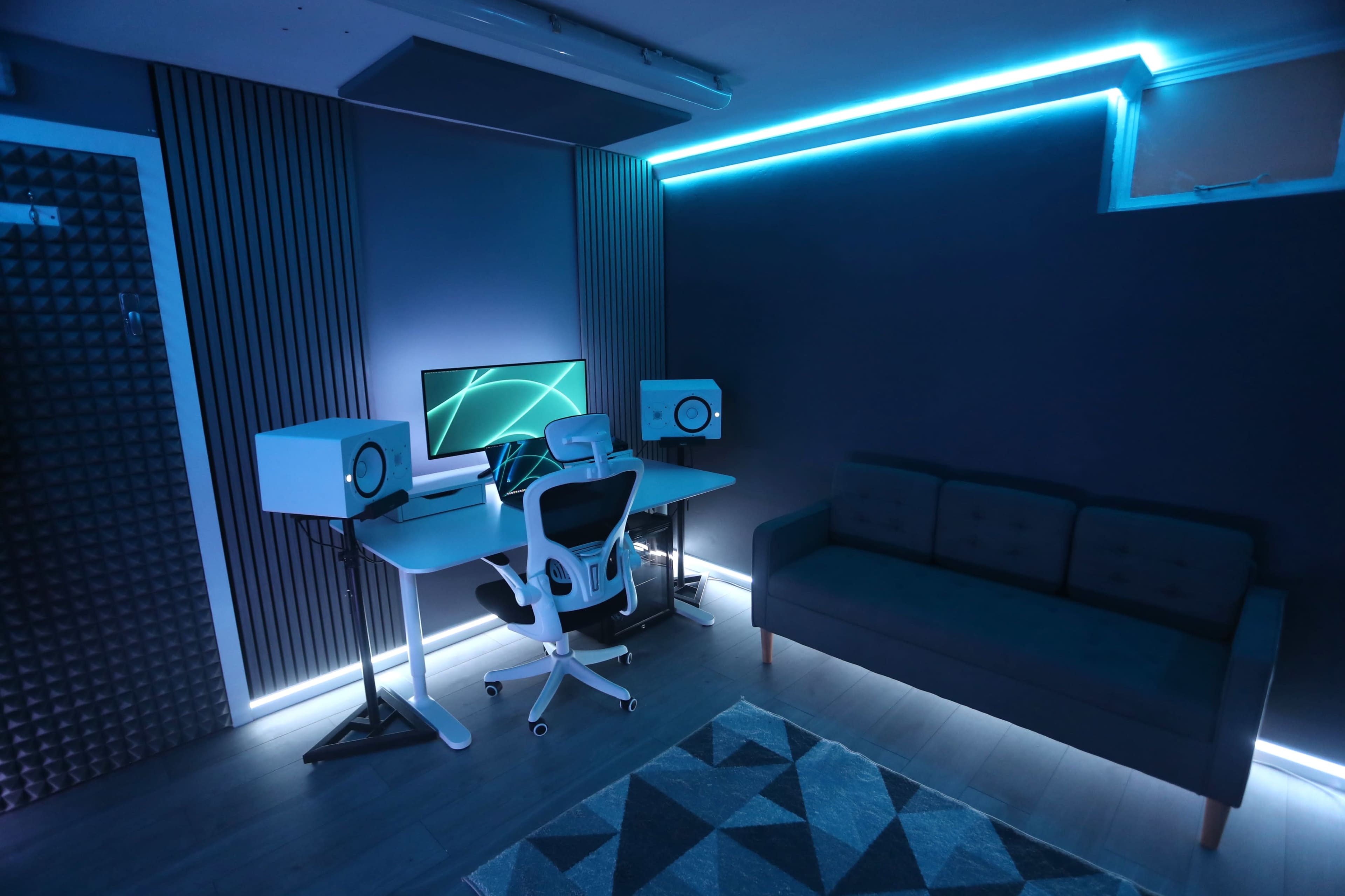 A modern workspace with a desk, computer monitor, studio monitors on either side, and a sofa, all illuminated by blue strip lighting.