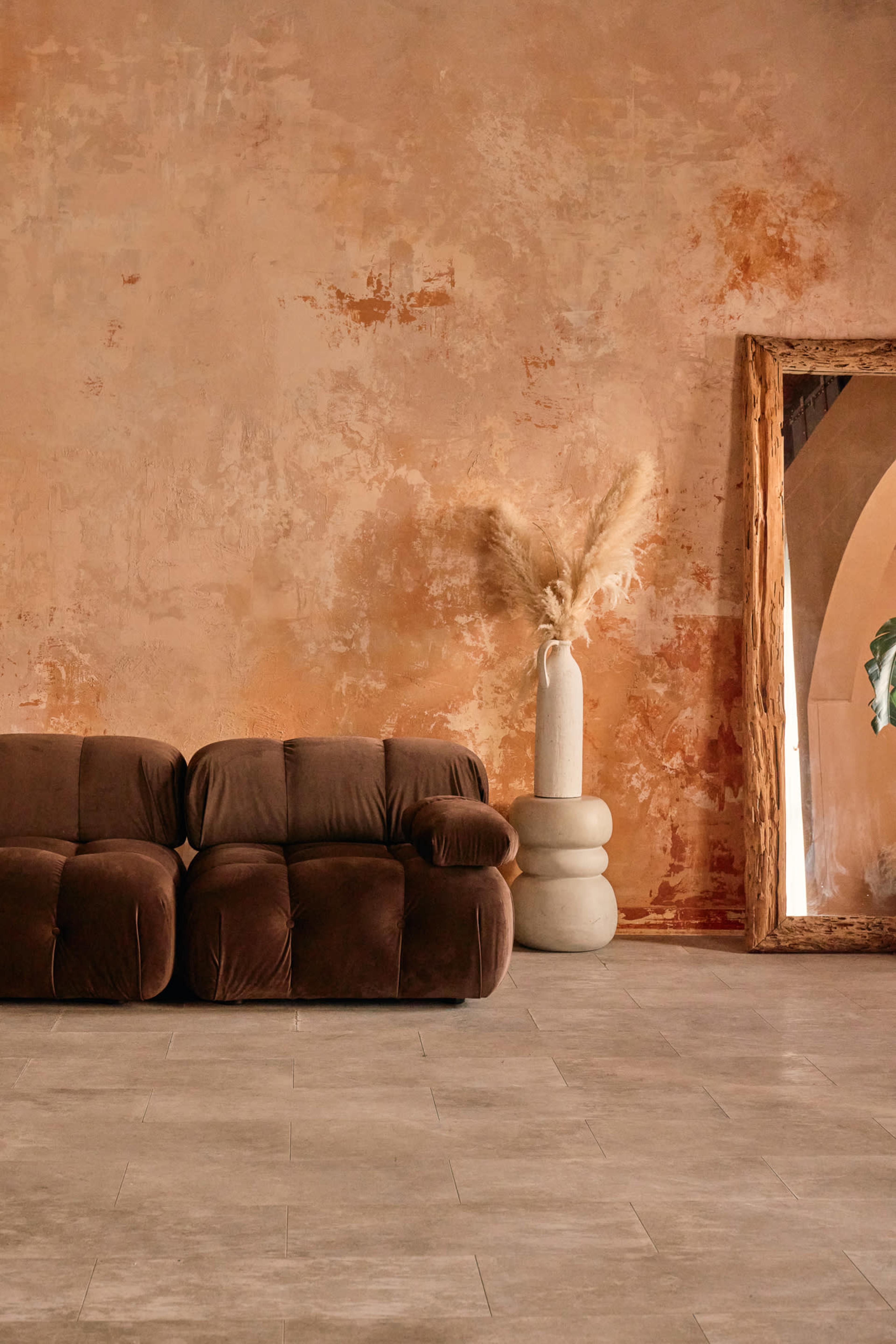 A brown, plush sofa sits against a textured, warm-colored wall, complemented by a tall vase with dried plants and a large, framed mirror nearby.