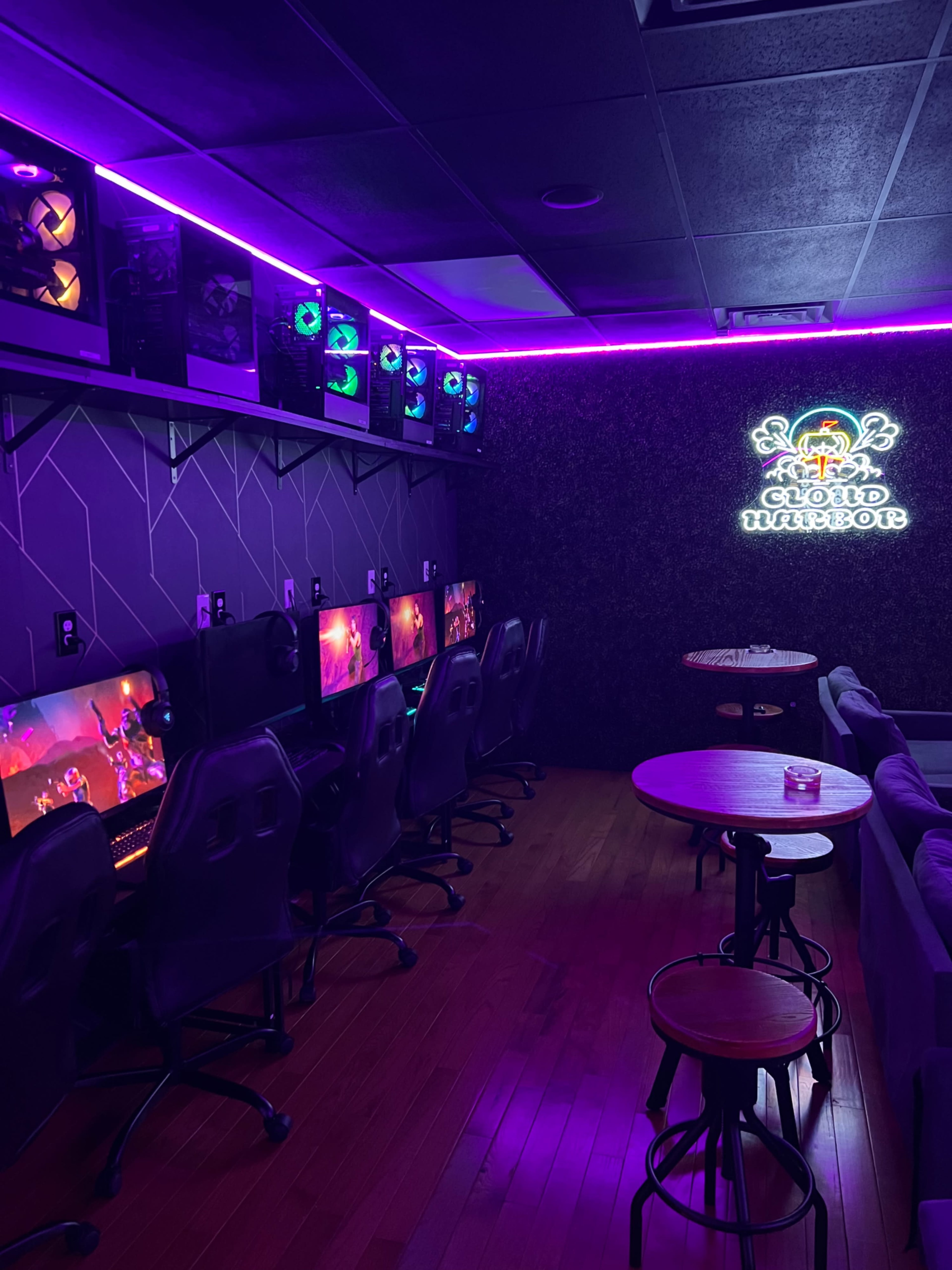 The image shows a gaming lounge with several computer stations along one wall, illuminated by purple lights, and a neon sign that reads "CLOUD WARRIORS."