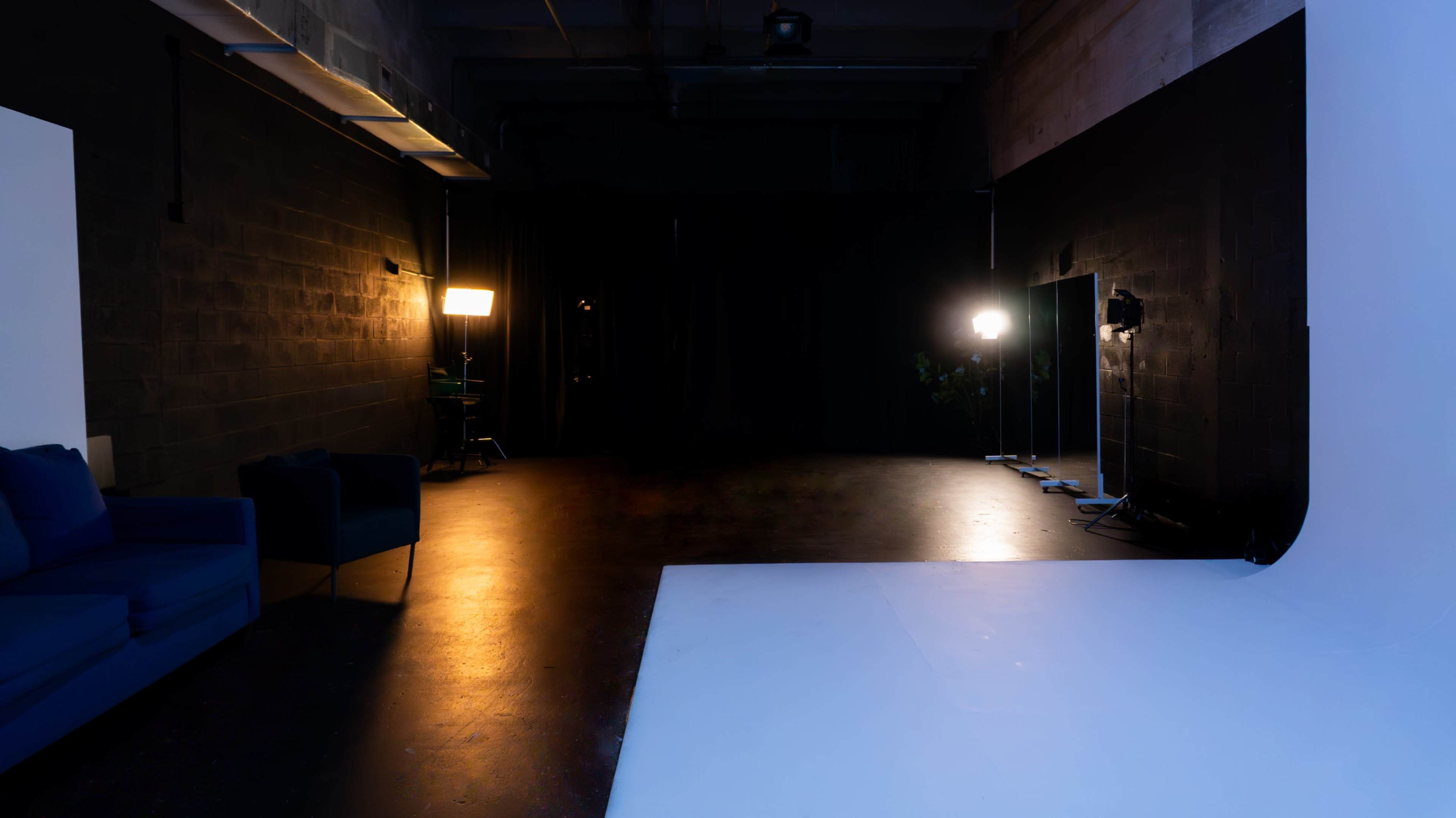 The image shows a dimly lit studio space with a white backdrop, a blue couch in the foreground, and lighting equipment positioned along the walls.