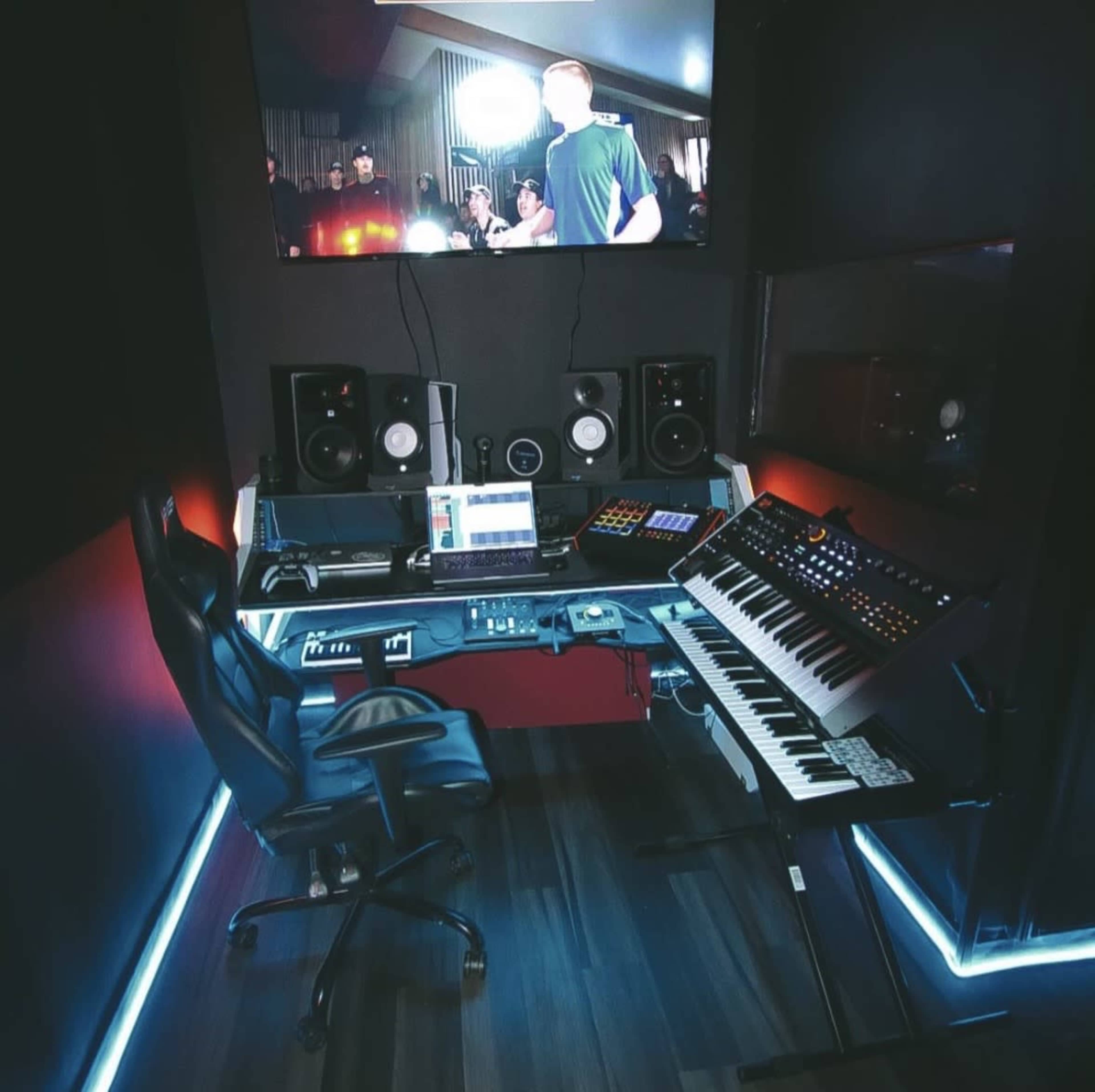The image shows a modern music production studio featuring a keyboard, audio equipment, and a large screen displaying a performance.
