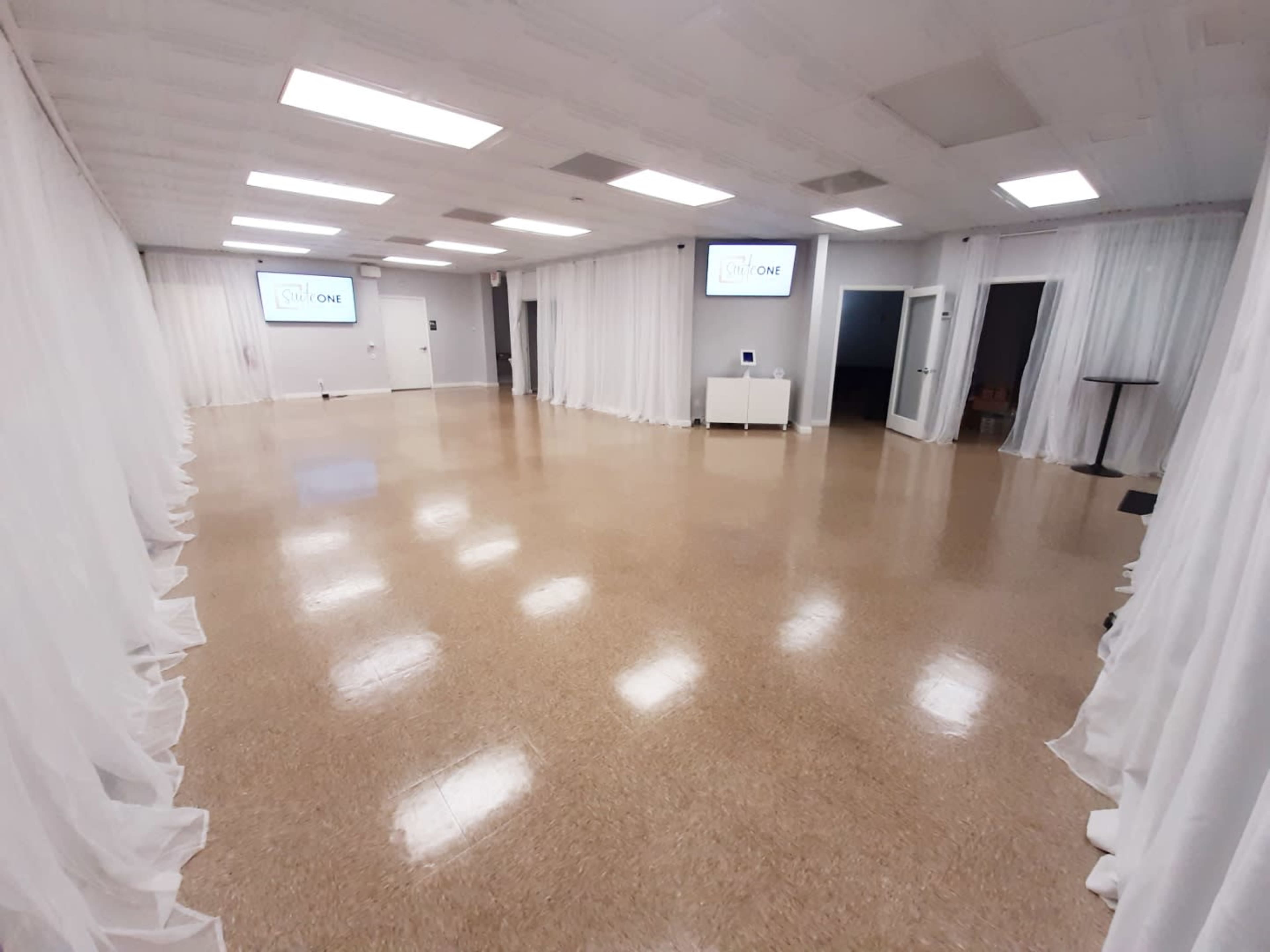 The image shows a spacious, empty room with polished flooring, white drapes along the sides, and two screens mounted on the walls.