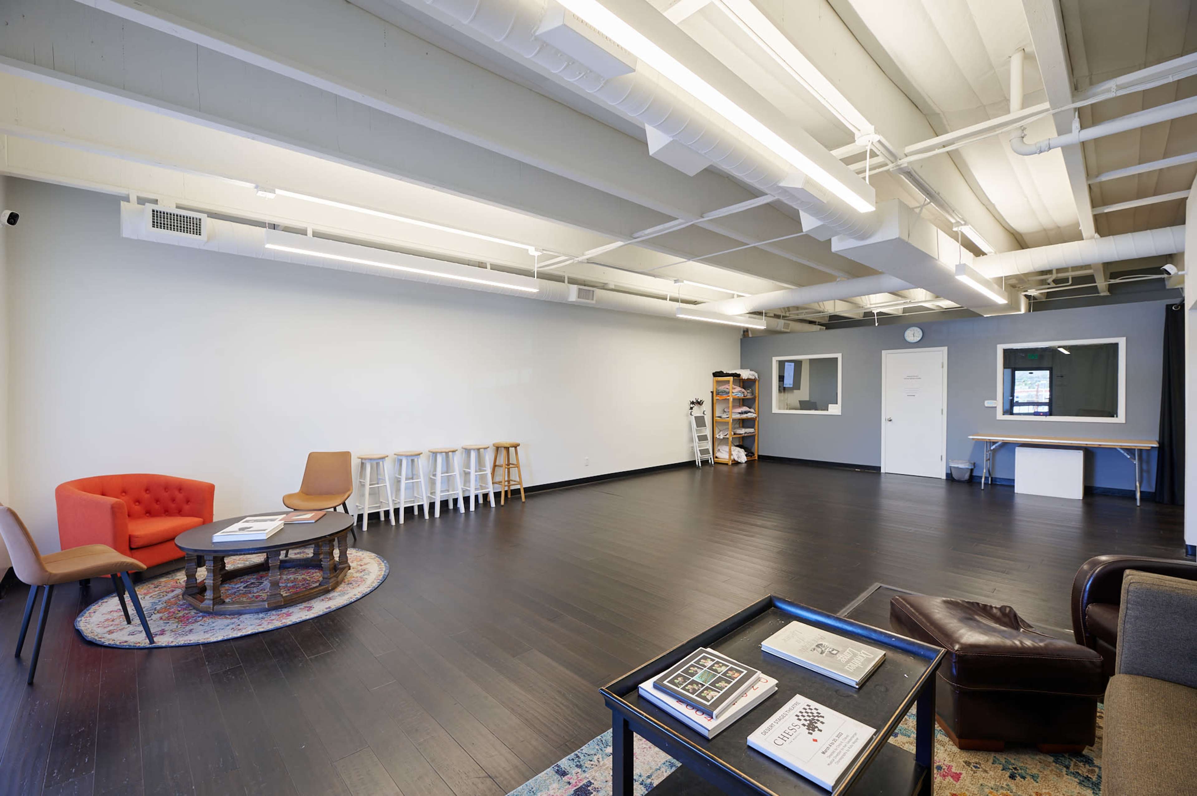 A spacious, minimalist room with dark wooden floors, a few scattered pieces of furniture including an orange chair, a brown sofa, and a round table.