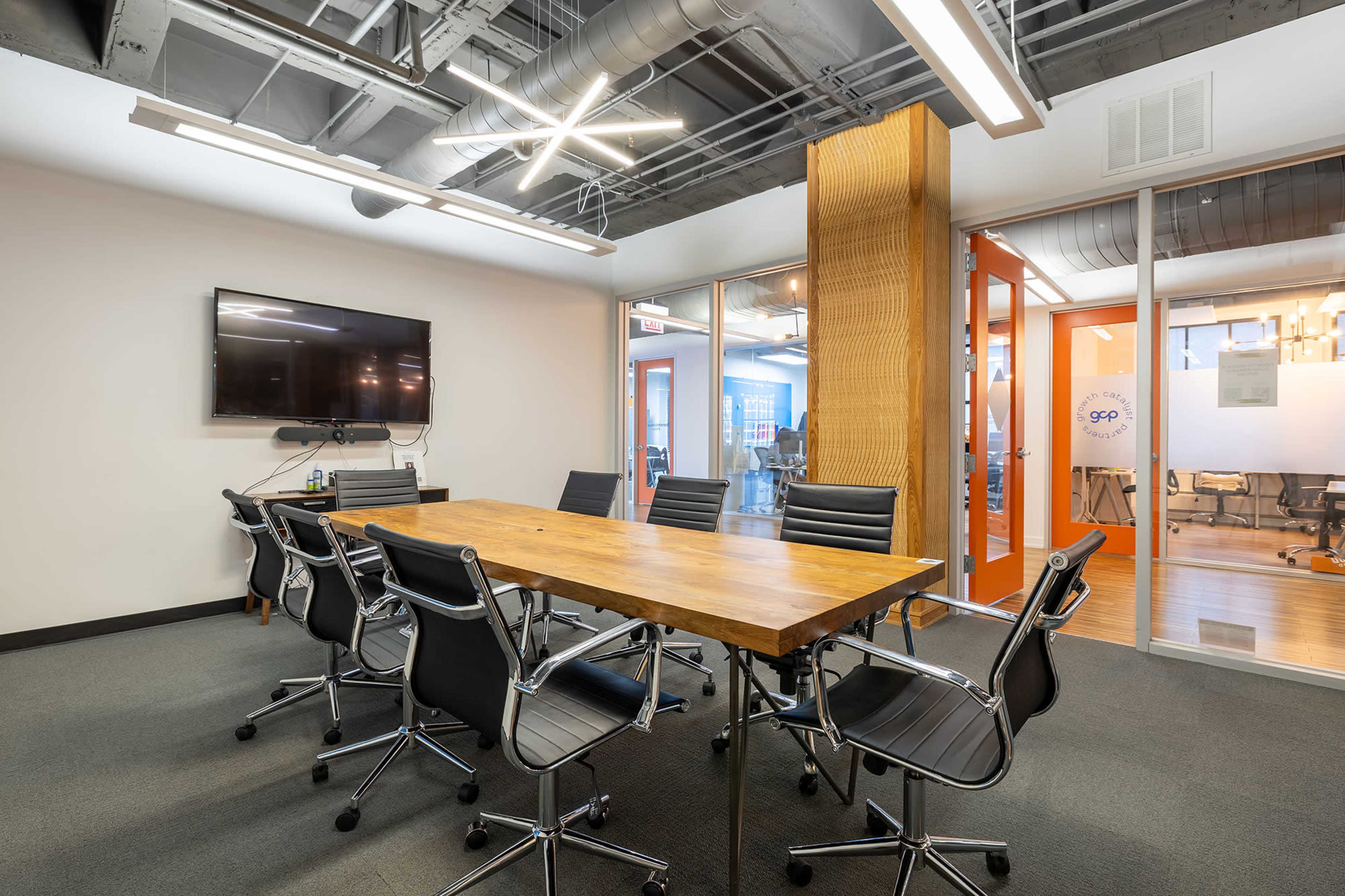 The image shows a conference room with a long wooden table surrounded by black rolling chairs, a wall-mounted television, and glass doors leading to an adjacent workspace.