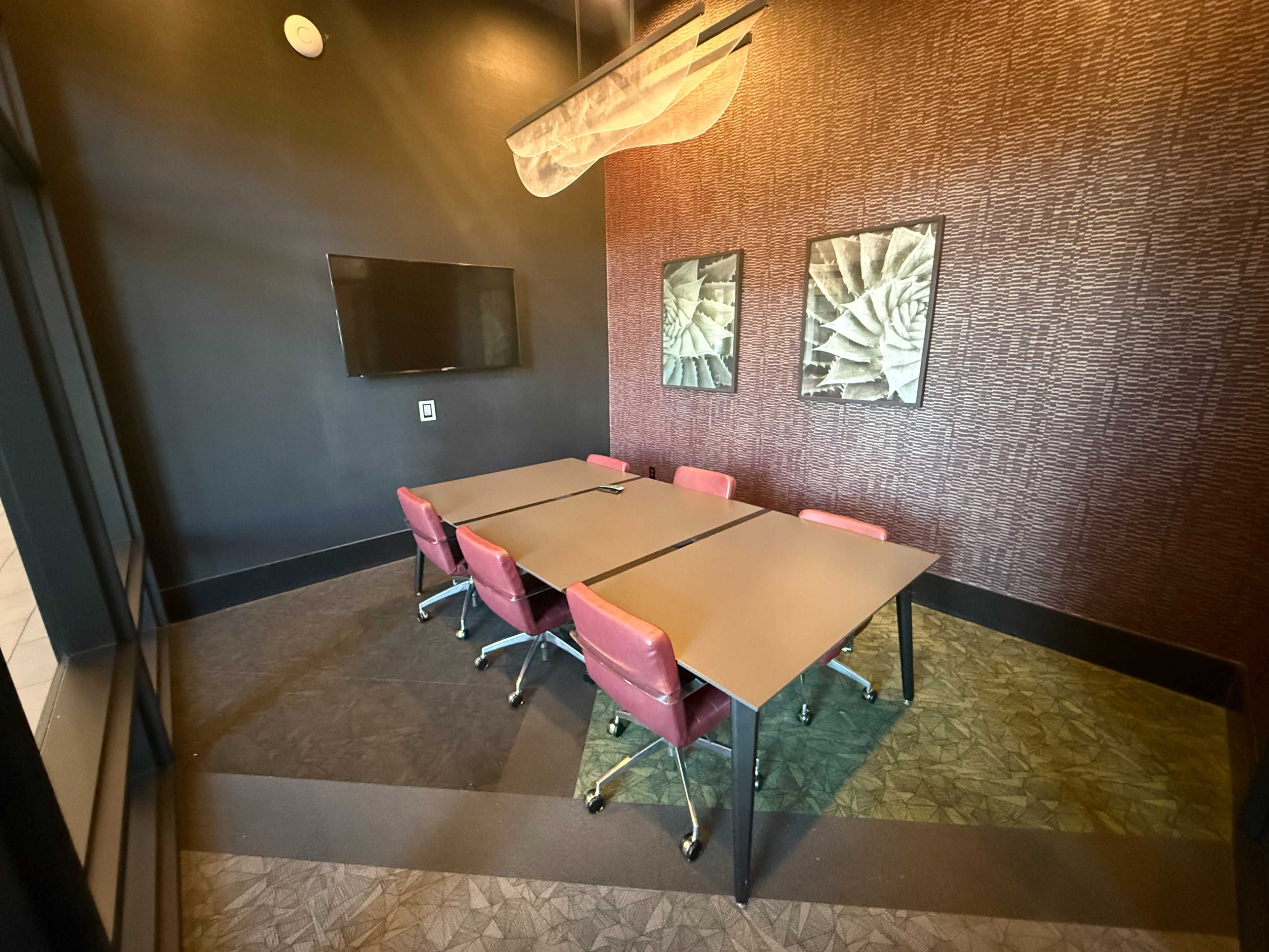 The image shows a small conference room featuring a rectangular table surrounded by red chairs, with two framed wall art pieces and a television mounted on the wall.