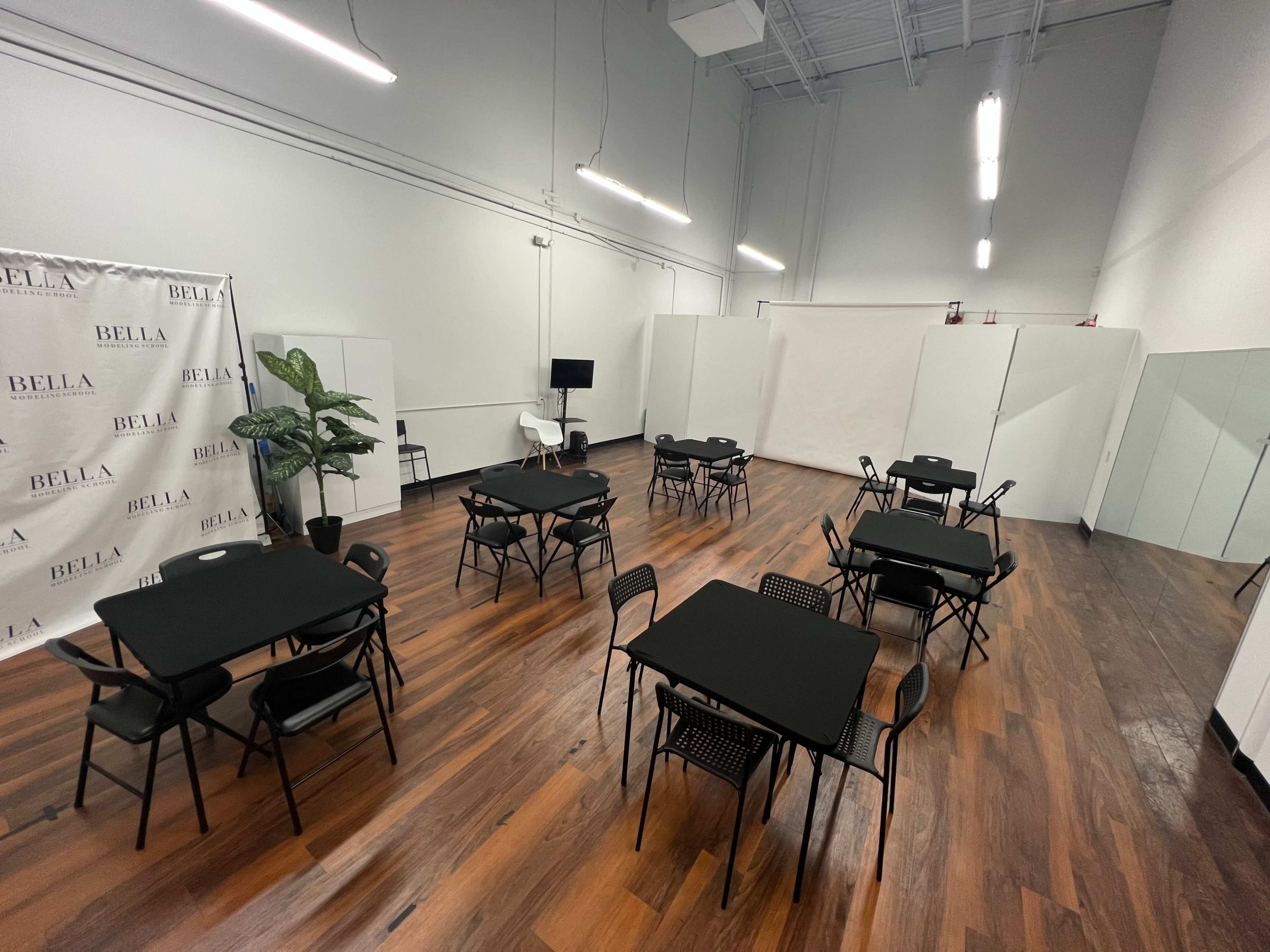 A spacious room with wooden floors, featuring several black tables and chairs arranged throughout, along with a plant and a background wall displaying the name "BELLA."