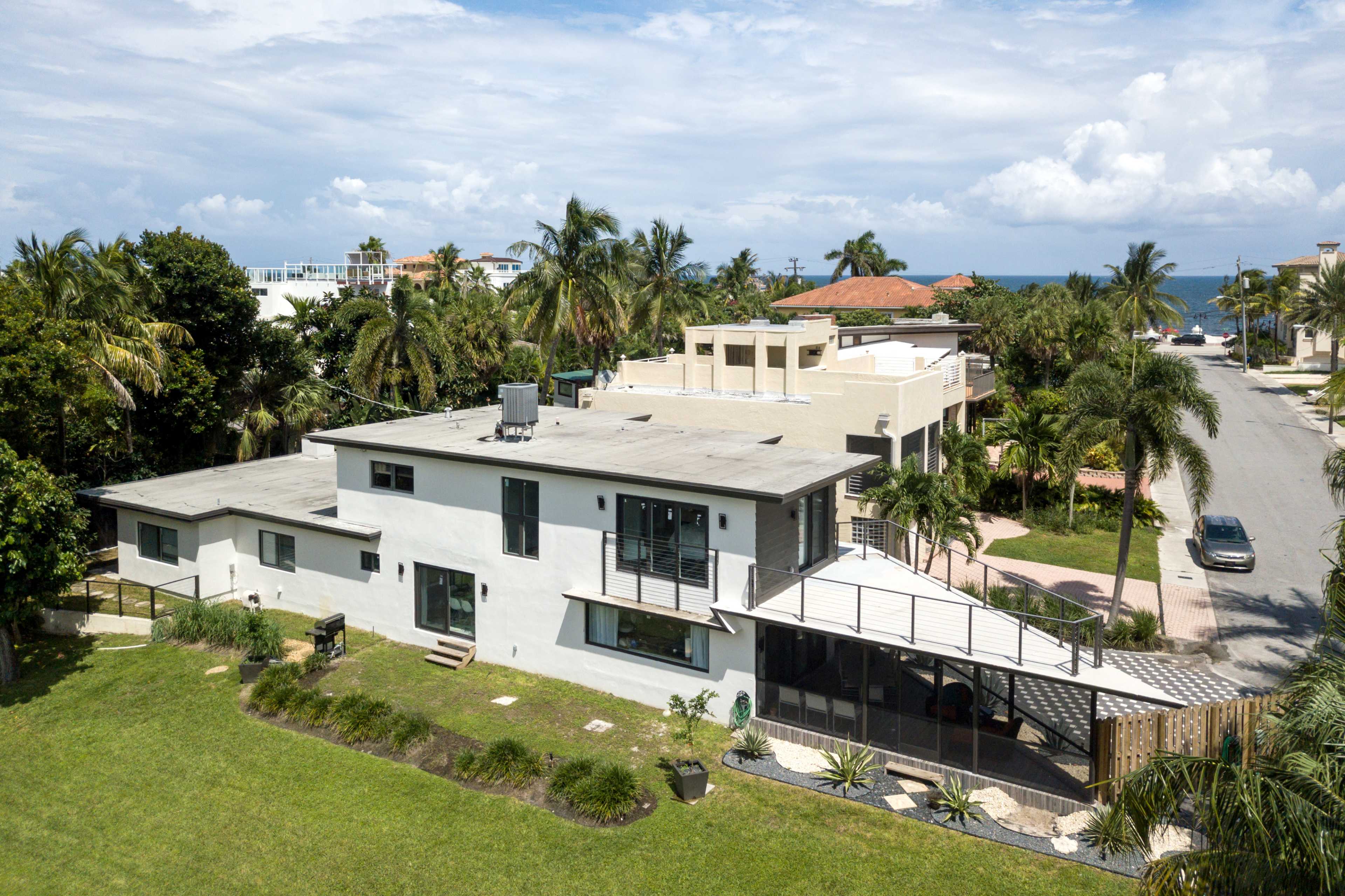 A modern two-story house with a flat roof and large windows is surrounded by greenery and palm trees, with a street and ocean visible in the background.