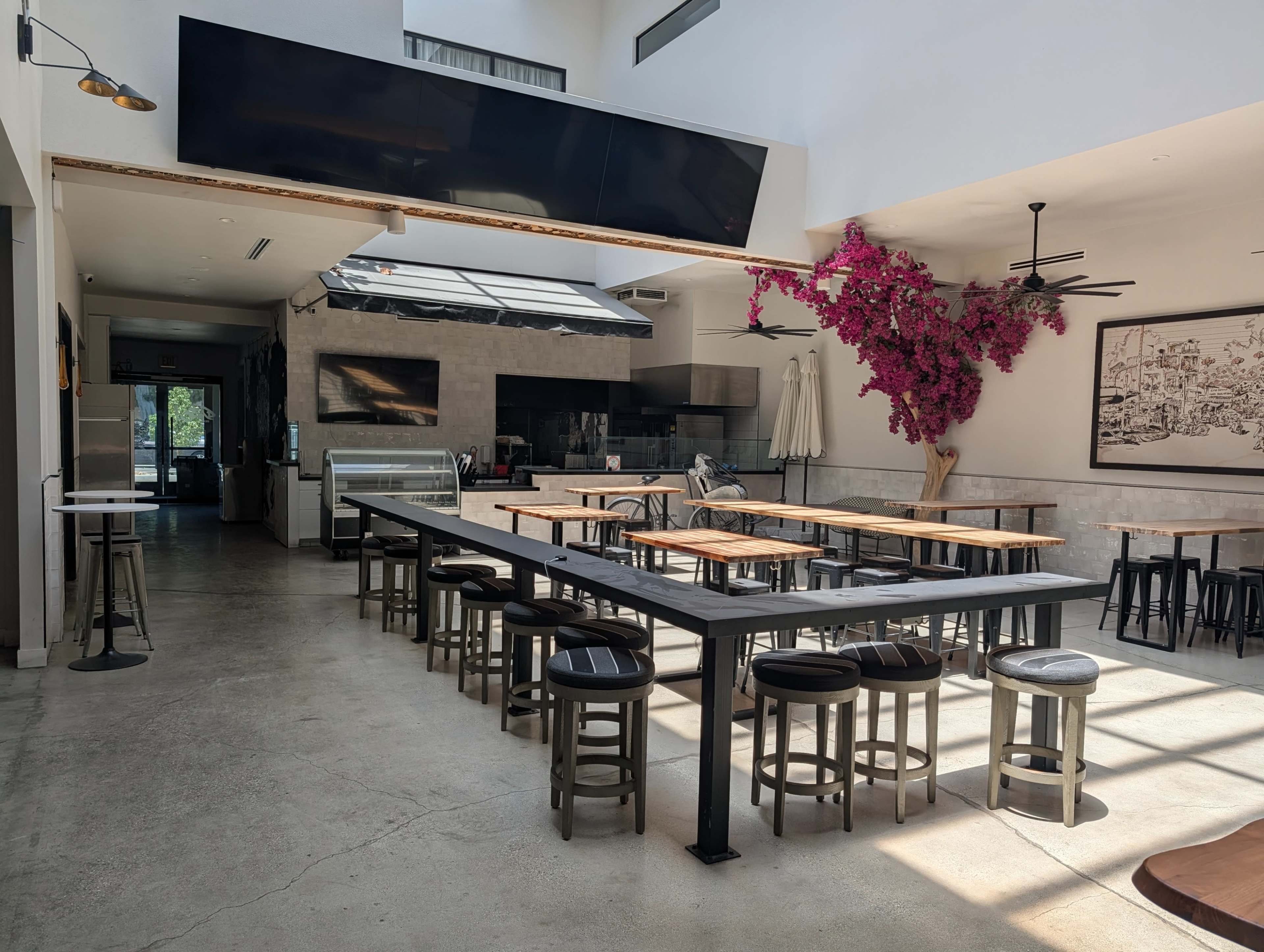 The image shows a spacious, modern restaurant interior with long tables, stools, and a prominent pink bougainvillea plant on the wall.