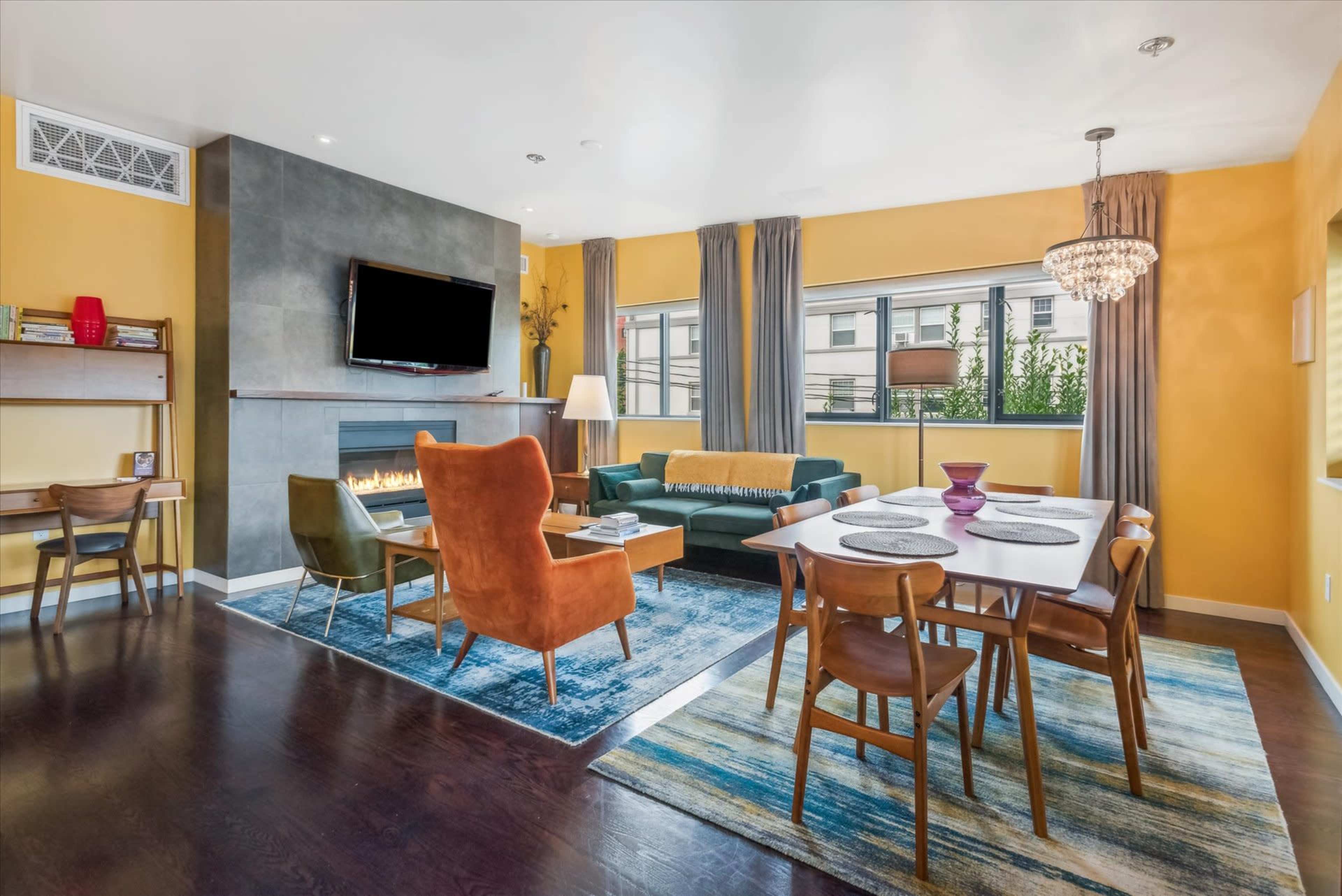 The image shows a modern living and dining area featuring a large window, a fireplace, a television, and a mix of furniture including a green sofa, orange armchair, and a wooden dining table with chairs.