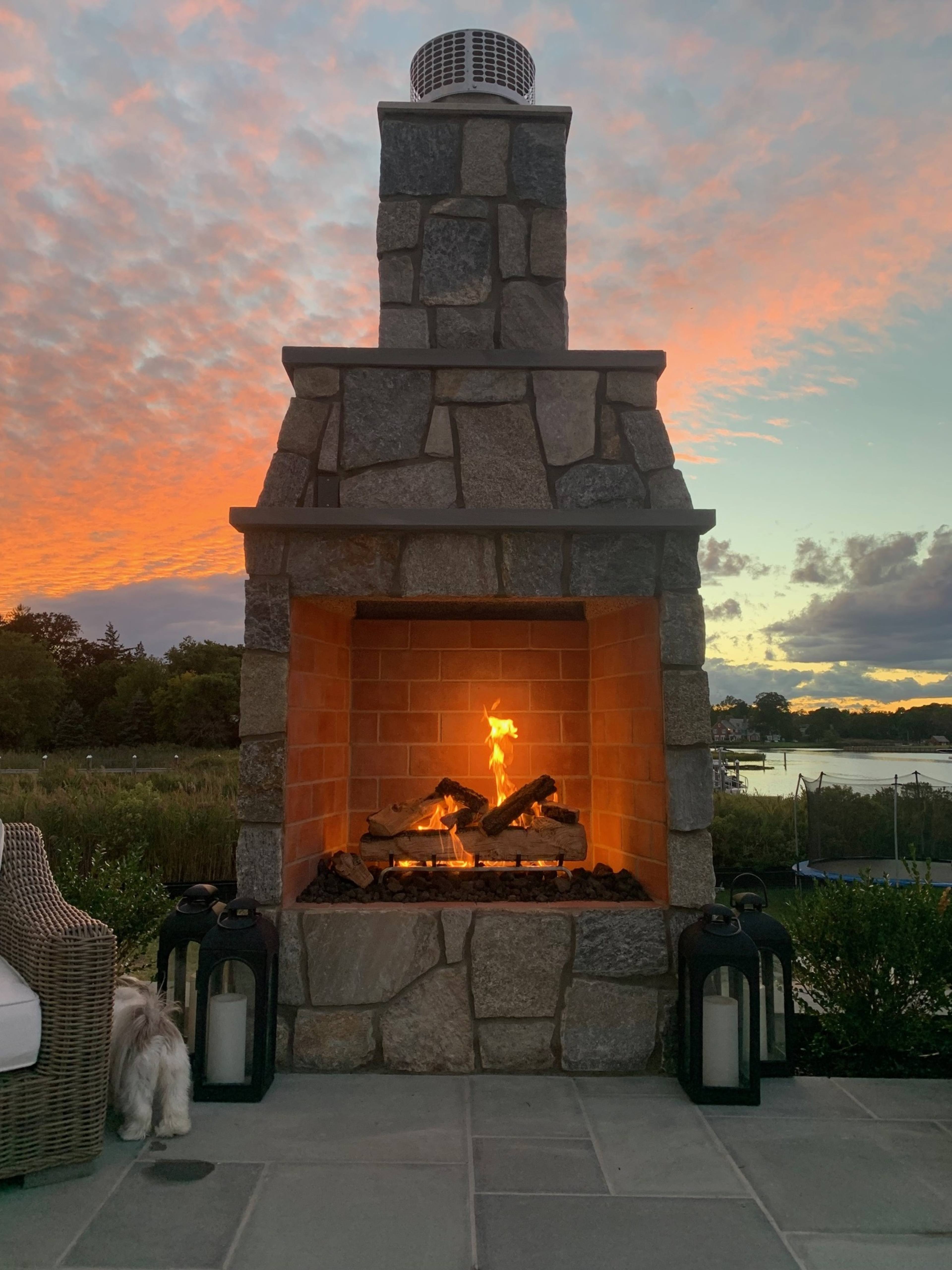 A stone outdoor fireplace with a lit flame is set against a vibrant sunset sky, near a body of water.