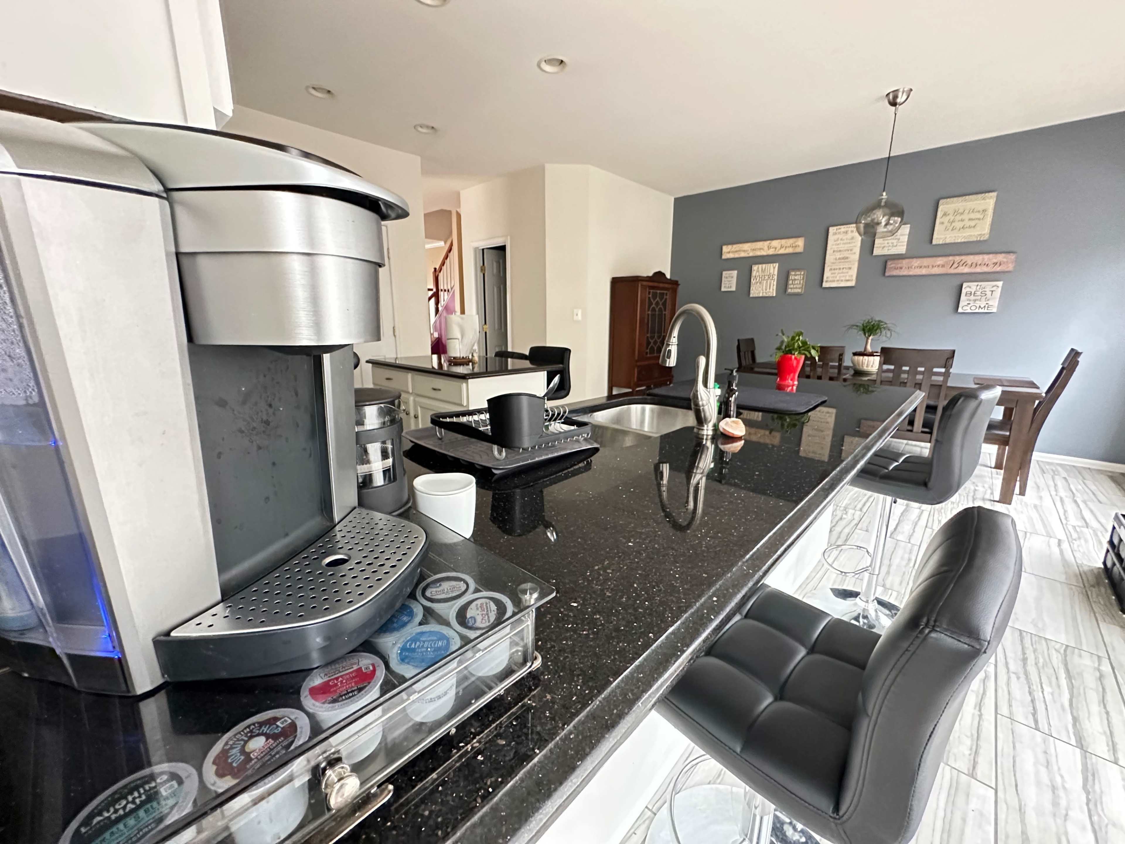 The kitchen features a coffee maker on the counter, a dining area with a table in the background, and a blend of dark and light-colored cabinetry.