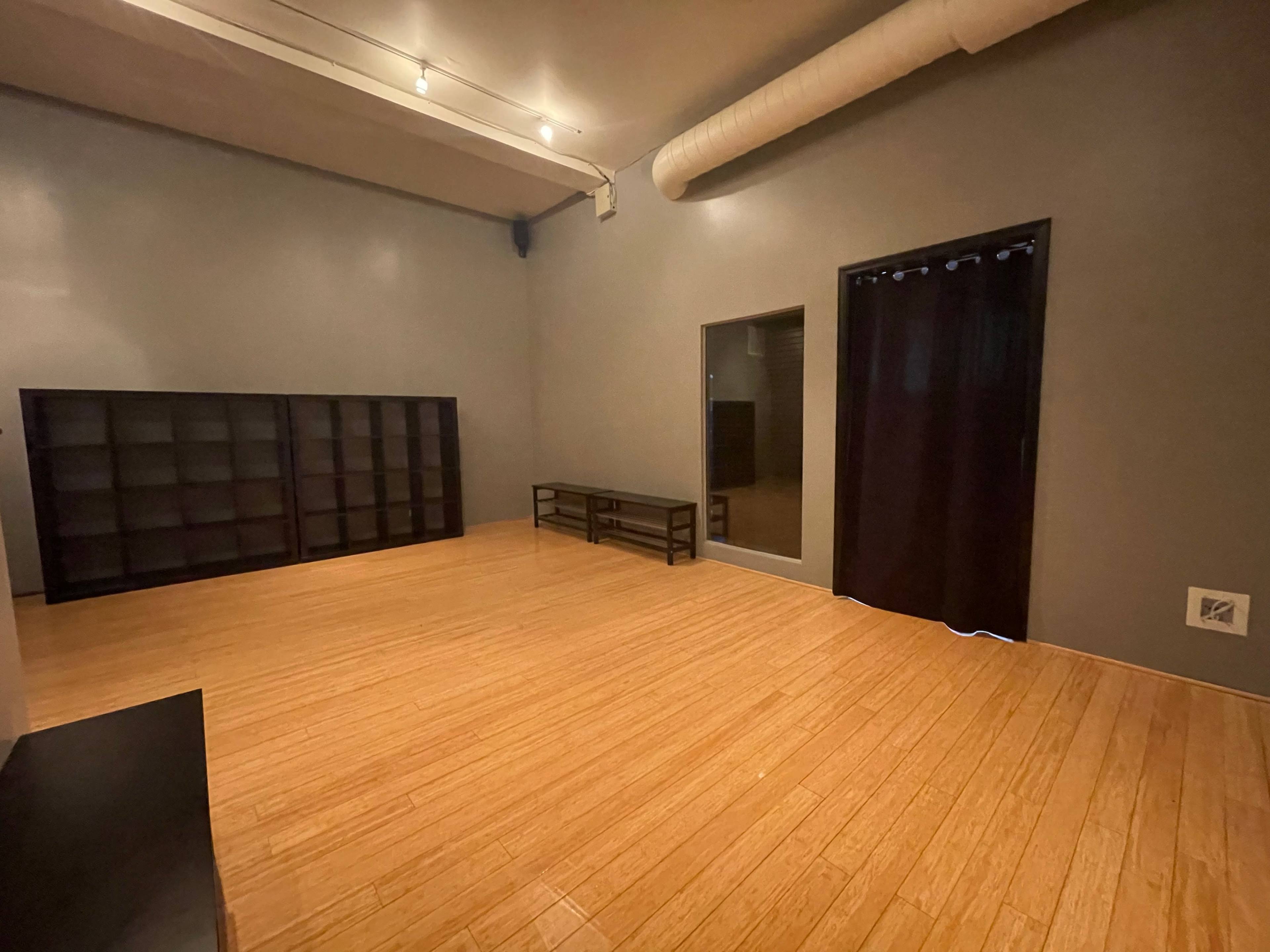 The image shows a spacious, empty room with wooden flooring, featuring a wall-mounted shelf, a low table, and a curtain-covered opening.