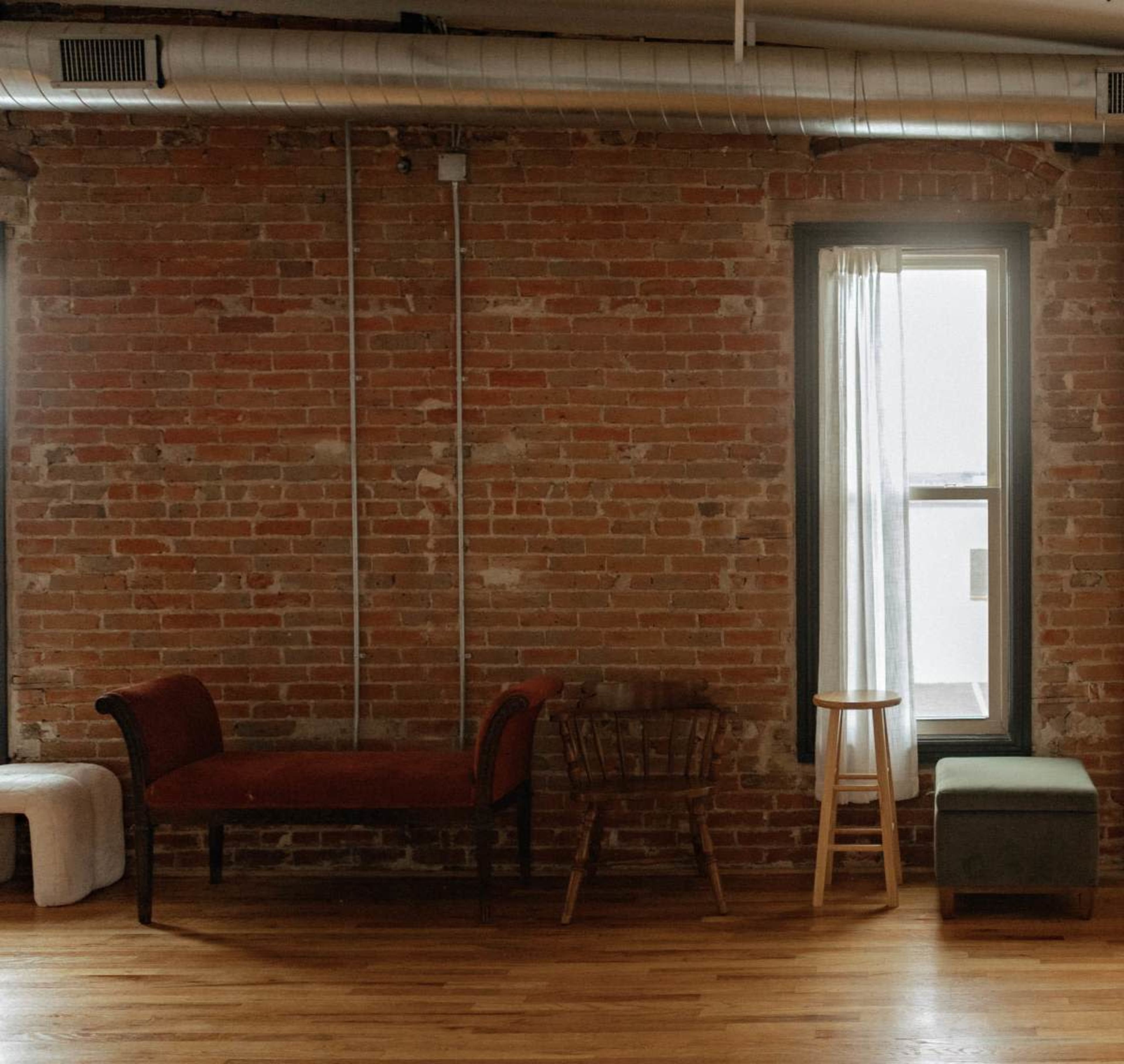 The image shows a room with exposed brick walls, featuring a sofa, a wooden chair, a stool, and a window with a sheer curtain.