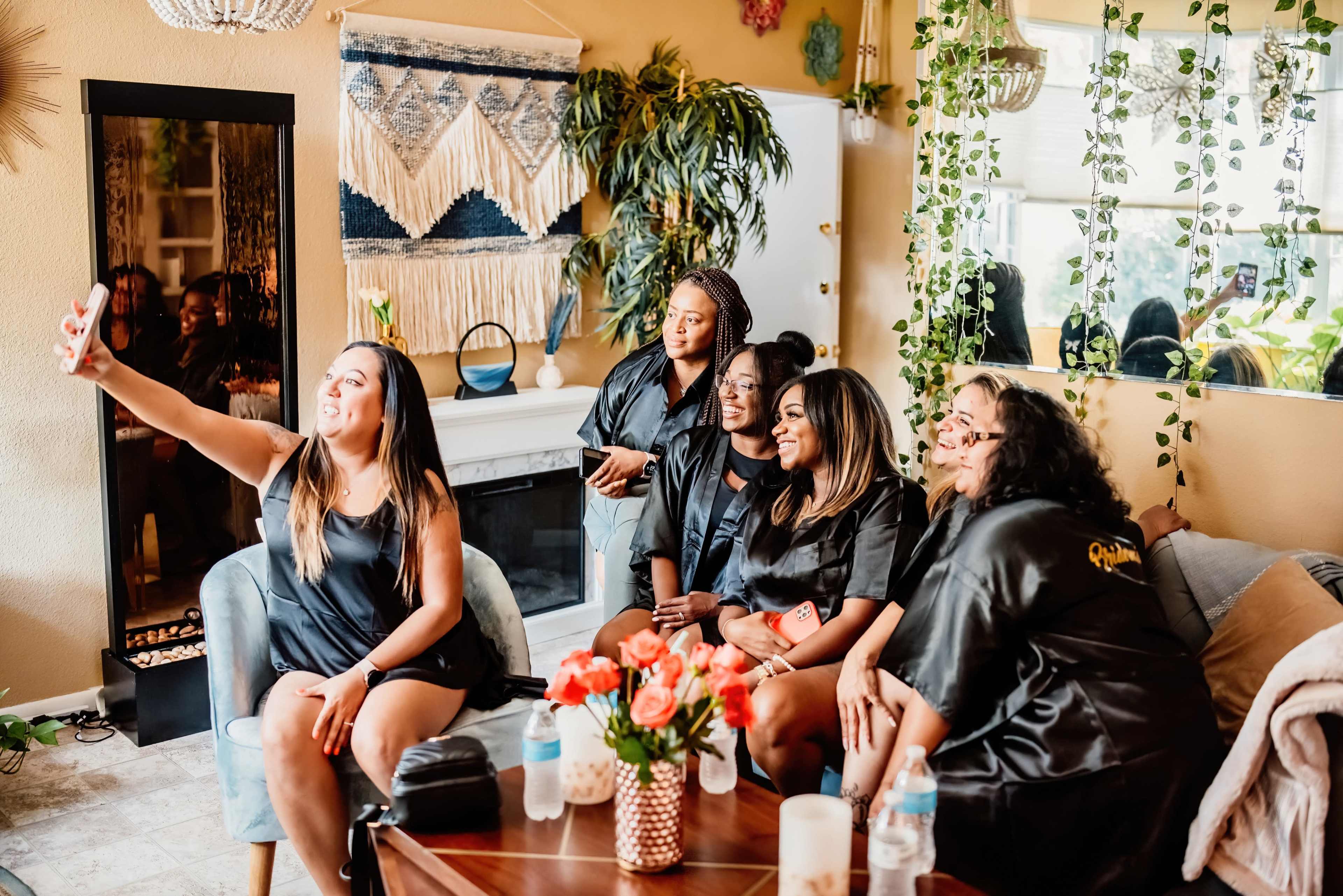 A group of six women in matching black robes pose together for a selfie in a cozy living room adorned with plants and decorative accents.