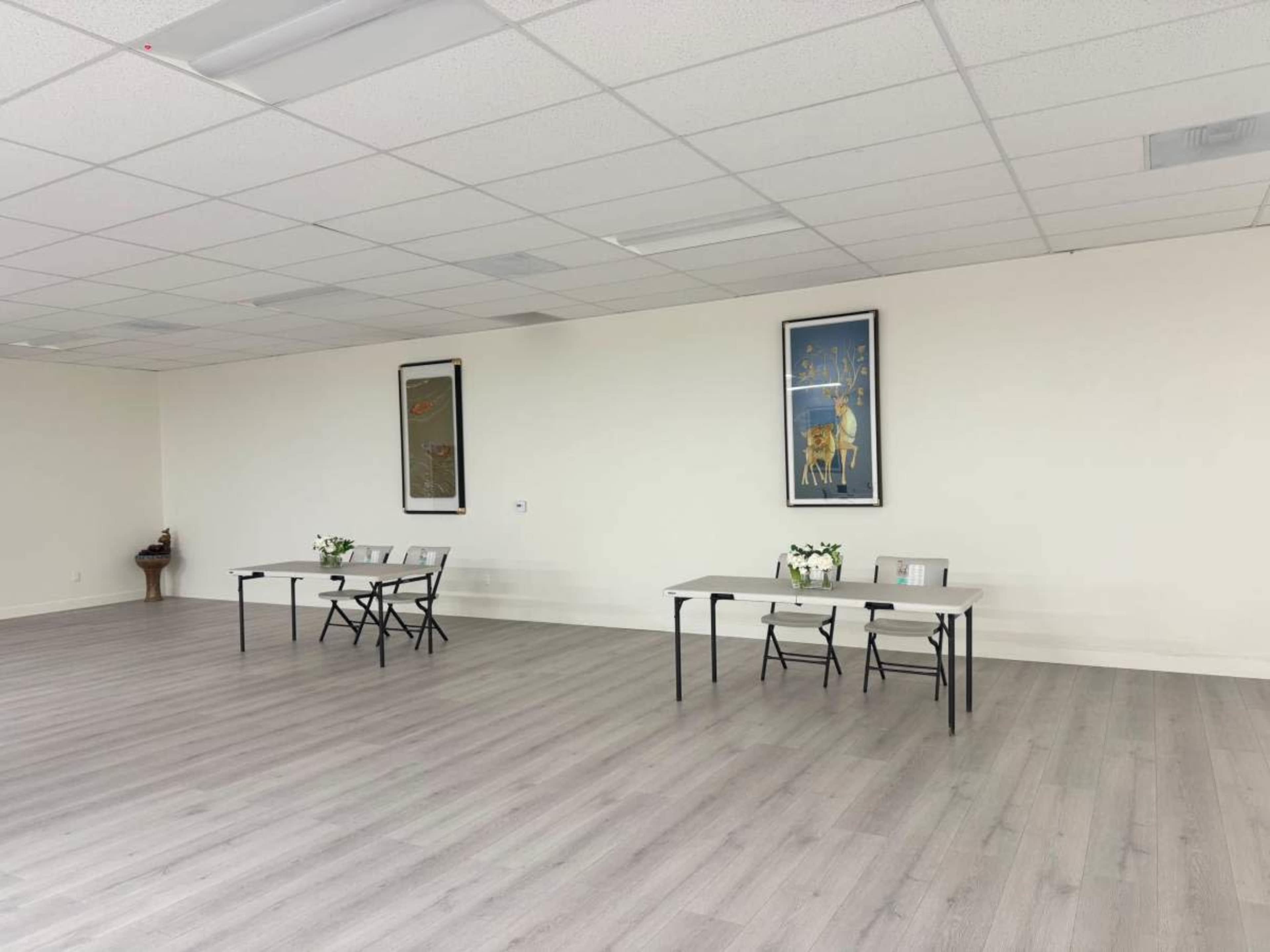 The image shows a spacious, empty room with two tables set up against a wall, each adorned with small flower arrangements and framed artwork.