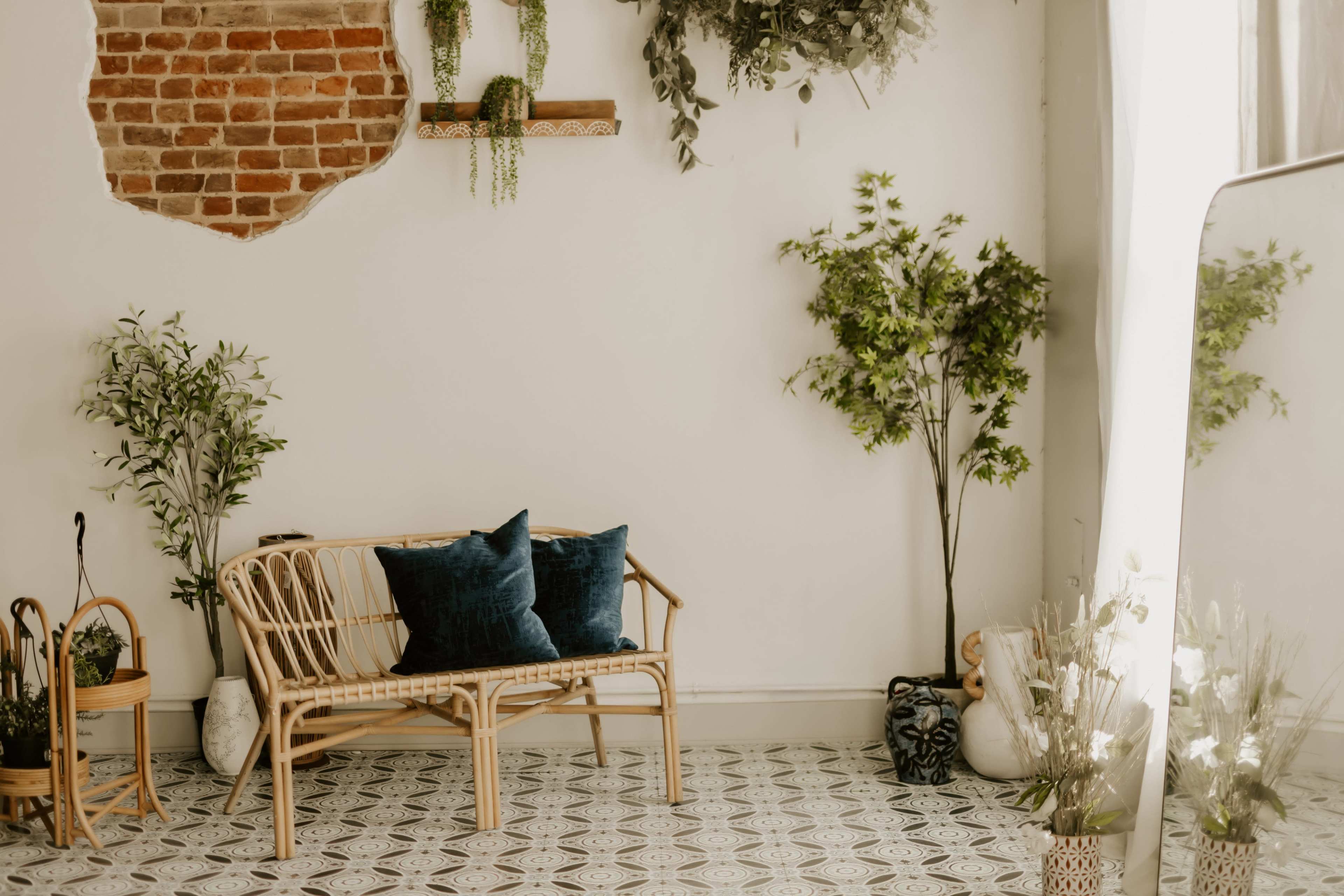 A rattan bench with blue cushions sits against a white wall adorned with greenery and a brick feature.