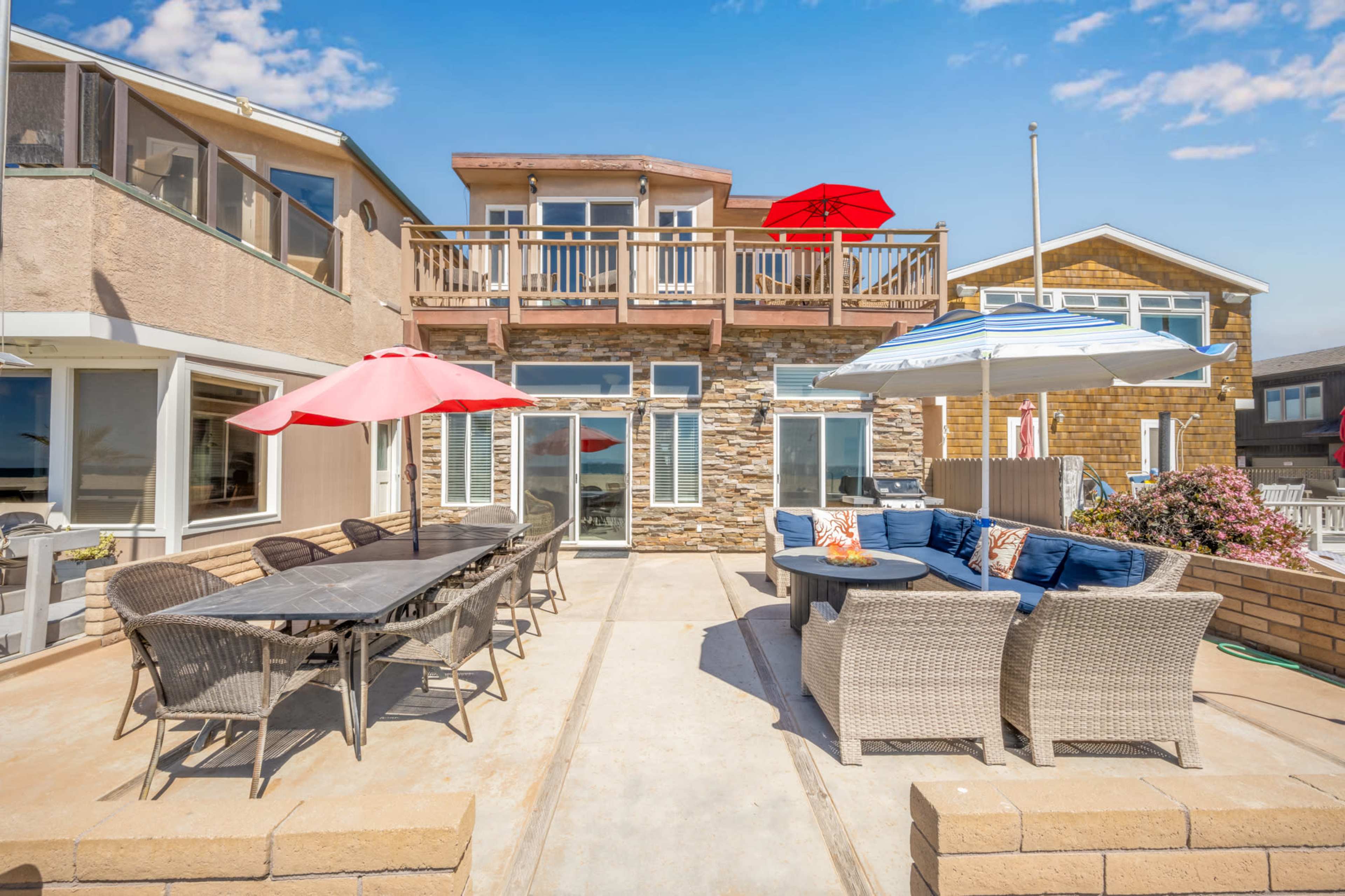 A patio area of a beach house featuring a large dining table, several lounge chairs, and umbrellas, surrounded by multiple buildings.