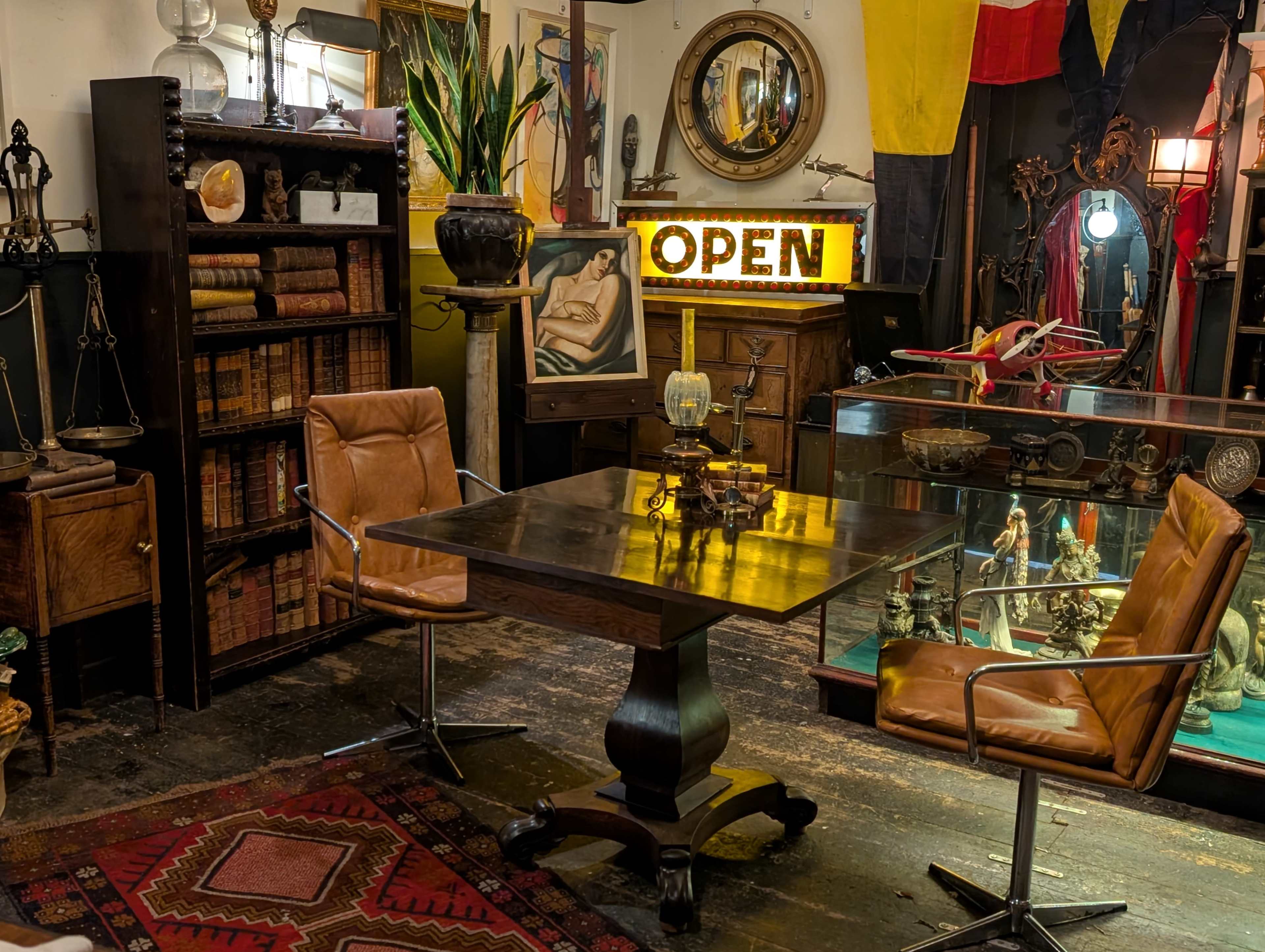 The image shows an antique shop interior featuring a wooden table surrounded by two chairs, with shelves of books, decorative items, and an illuminated "OPEN" sign in the background.