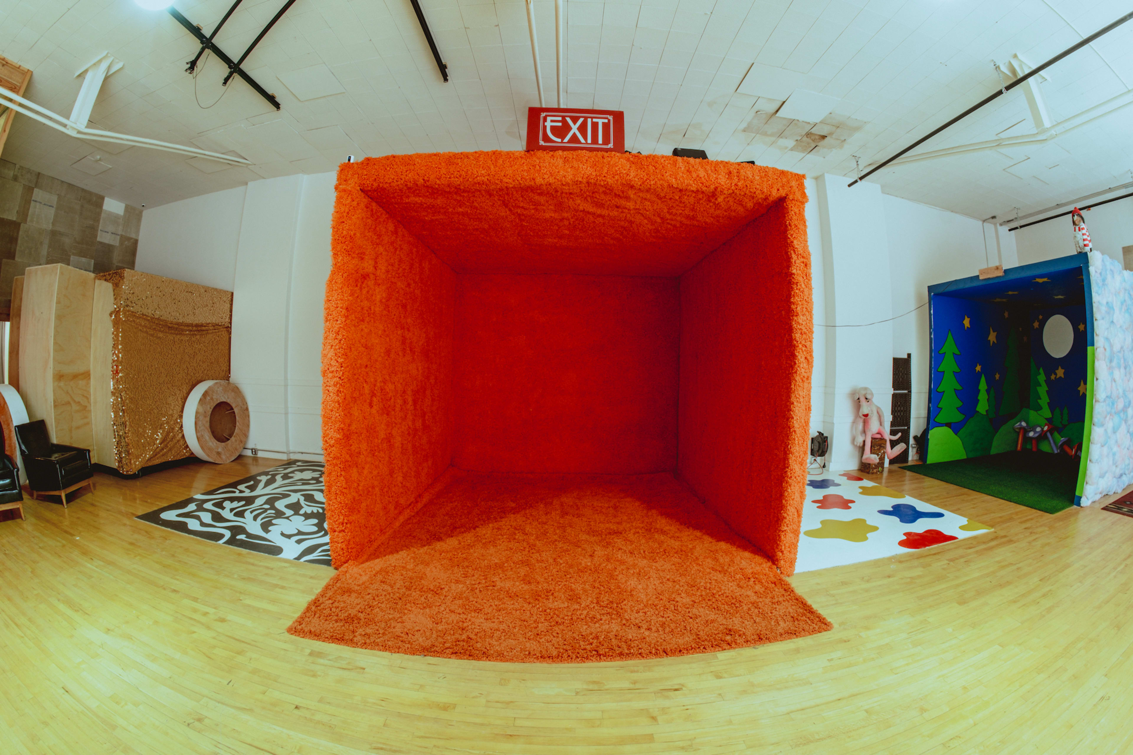The image shows a large, empty orange room with a textured carpet, featuring an exit sign above and colorful floor decorations nearby.