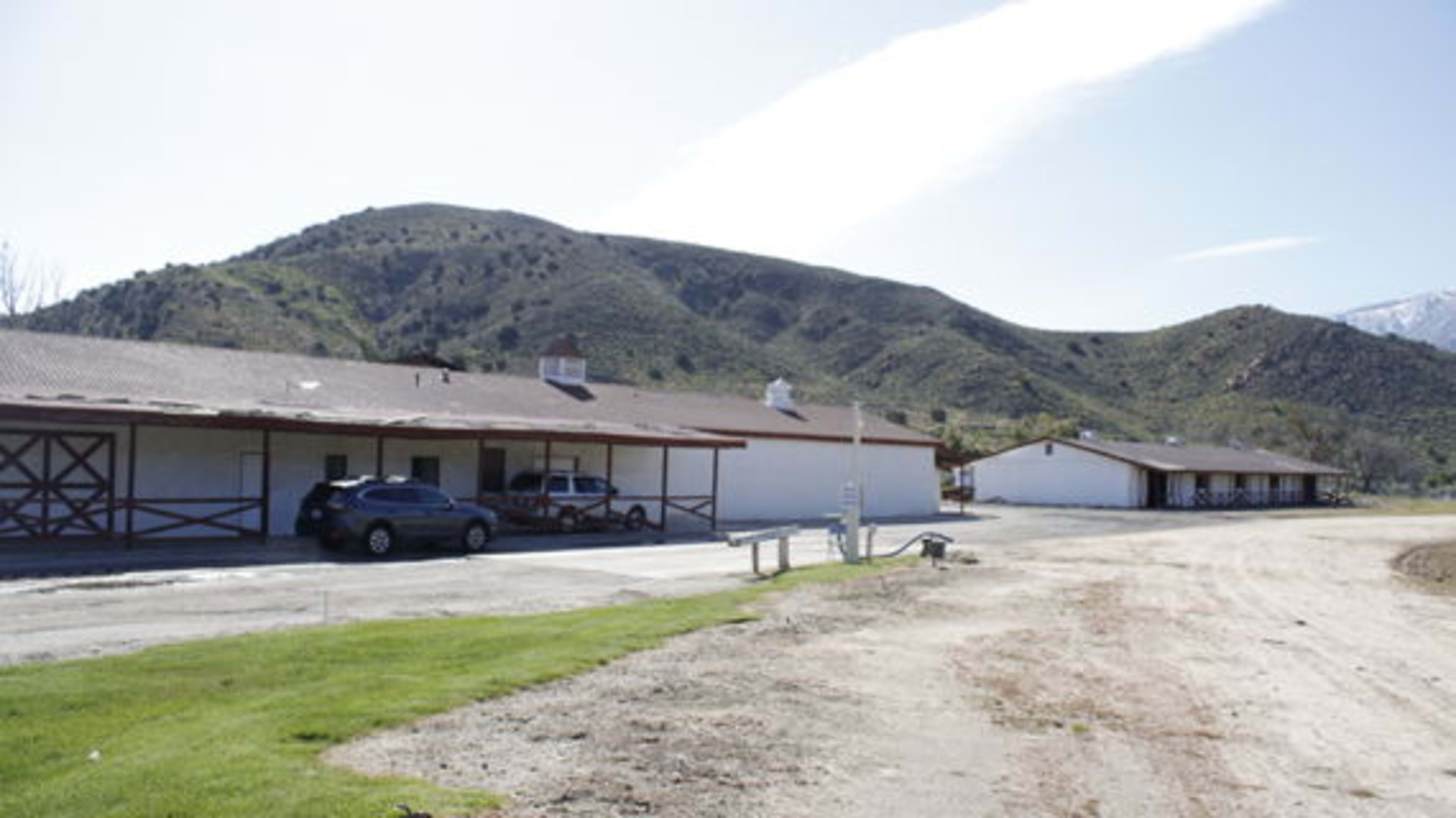 A rural landscape with a row of buildings adjacent to a dirt road, set against a backdrop of green hills.