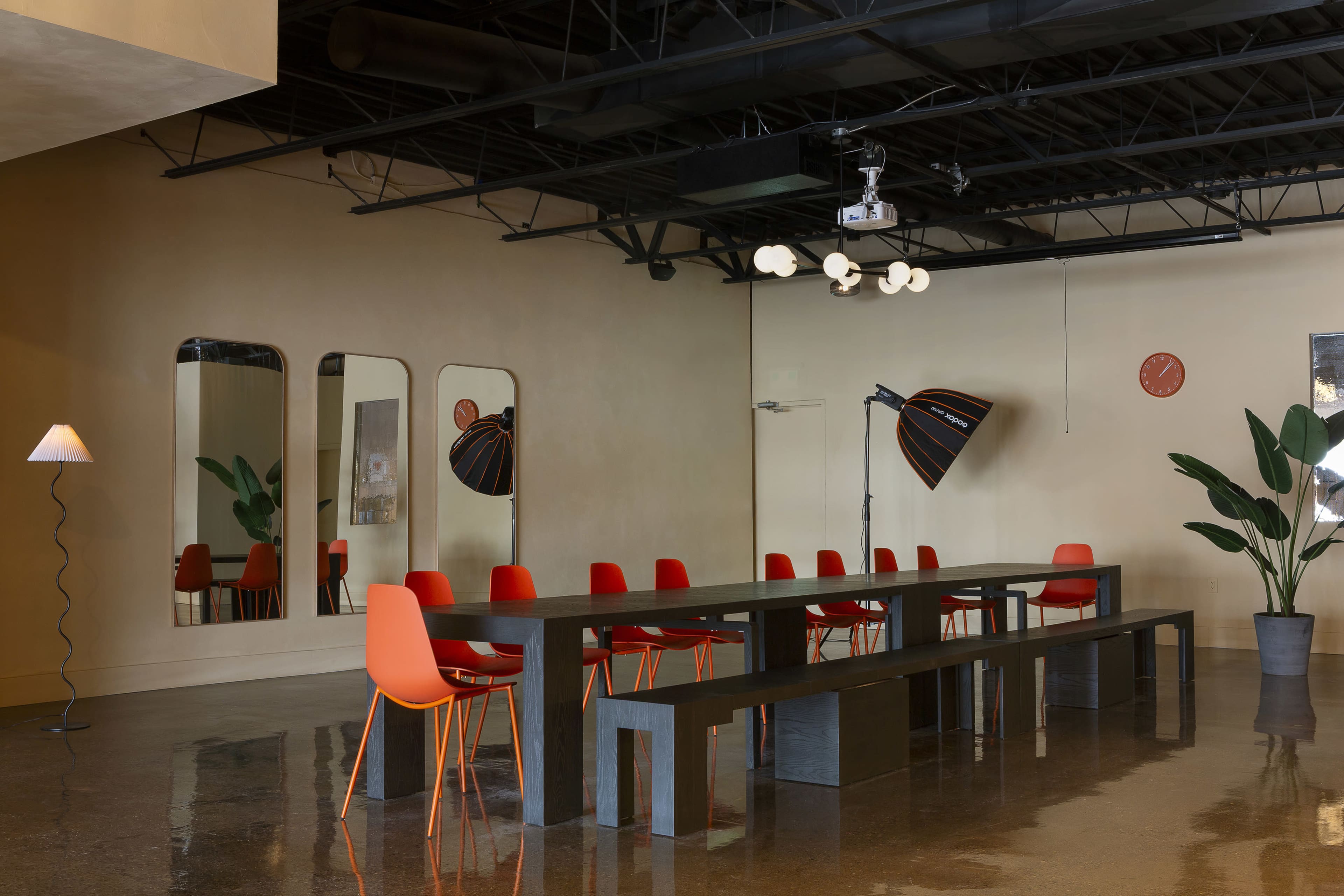 A spacious modern room features a long dark table surrounded by red chairs, with large mirrors and stylish lighting fixtures illuminating the space.