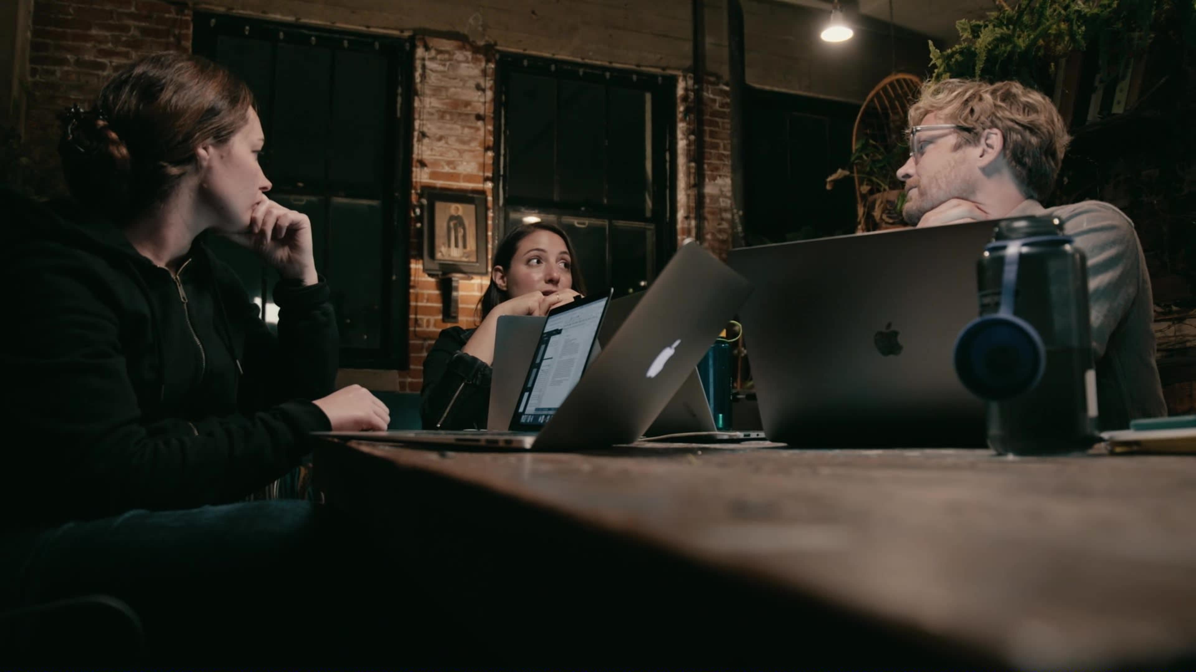 Three people engage in conversation around a wooden table, with laptops open in front of them in a softly lit room featuring brick walls.