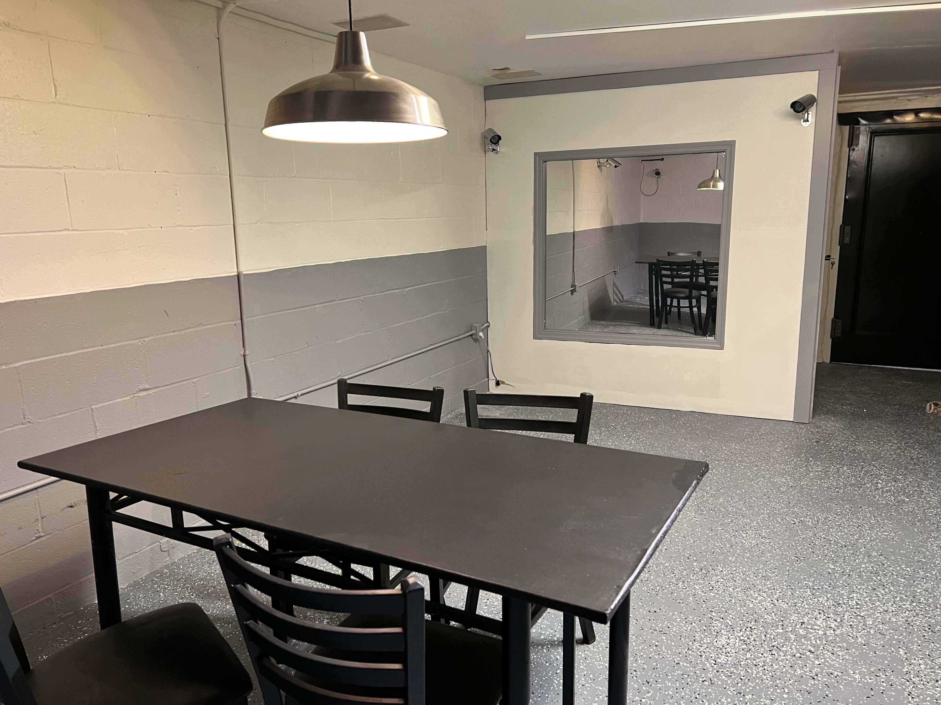 A simple room with a black table and chairs, featuring a large mirror on one wall and industrial-style lighting.