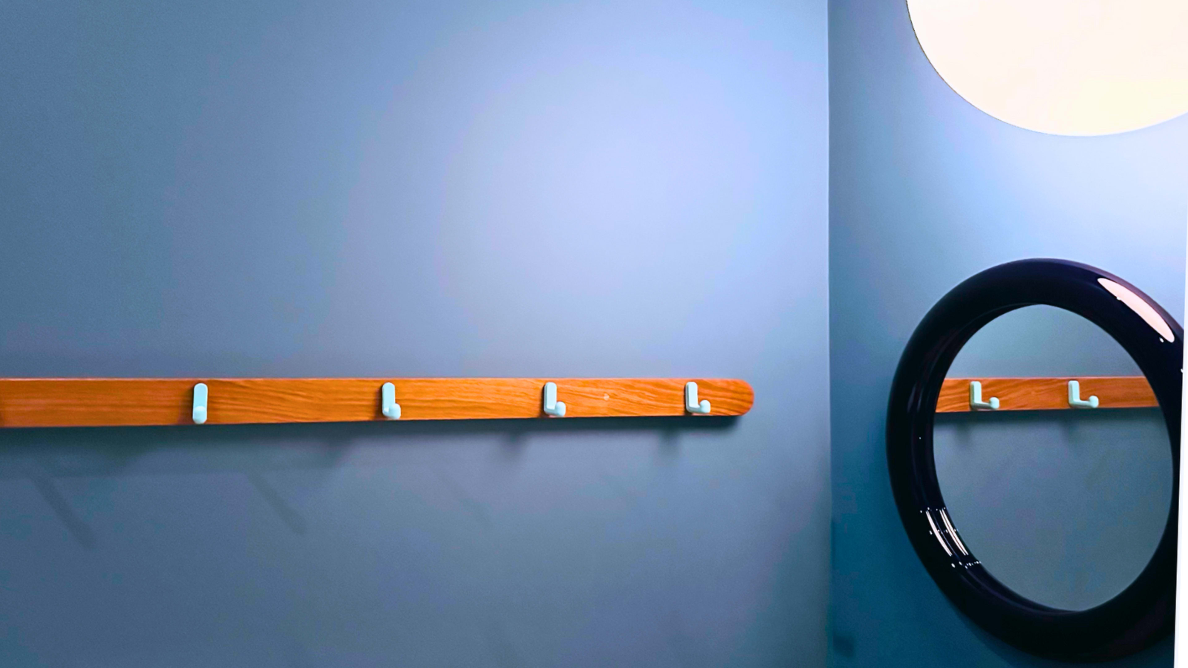 The image shows a minimalist corner with a wooden wall rack featuring several hooks and a circular mirror mounted on a light blue wall.