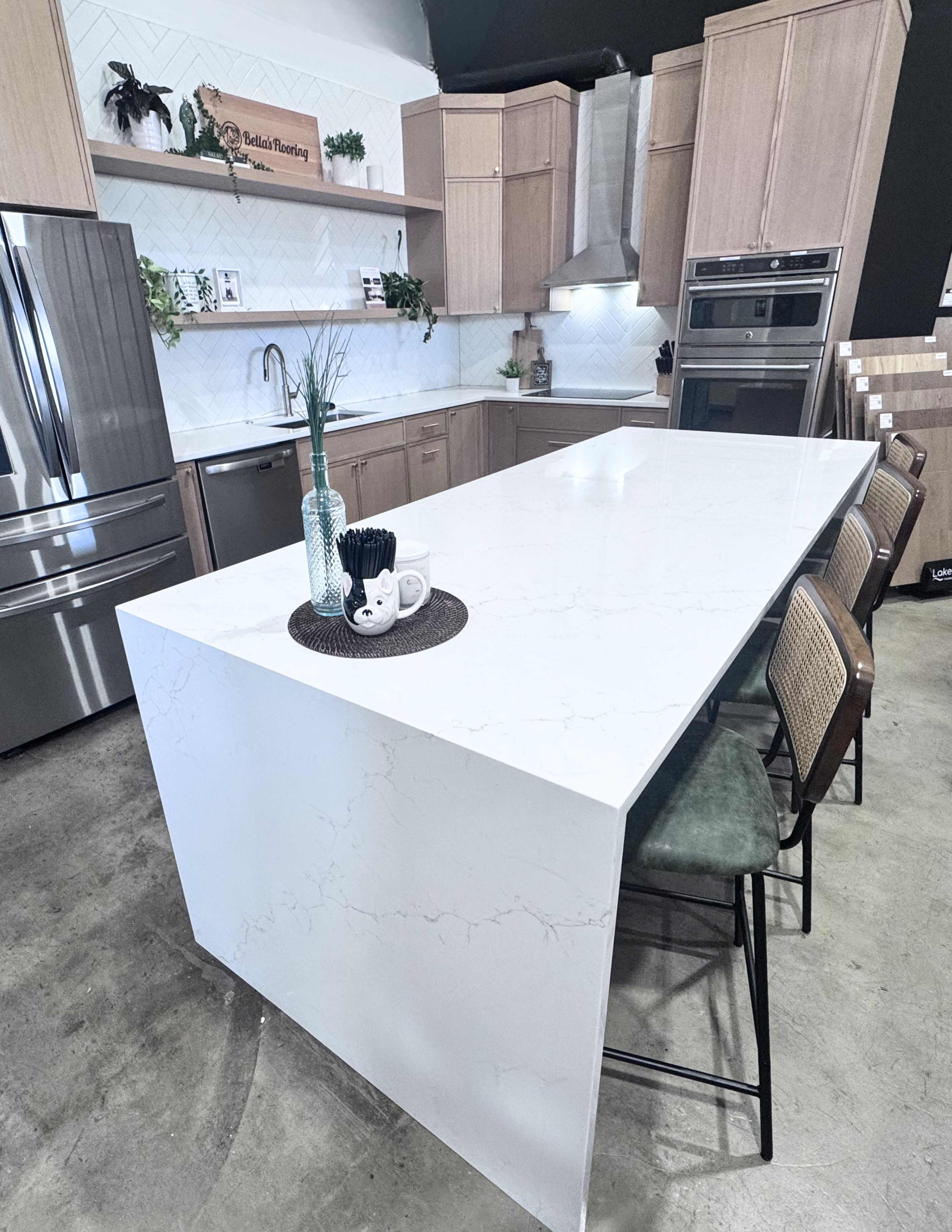 The image shows a modern kitchen with light wood cabinets, a large white marble island, and stainless steel appliances.