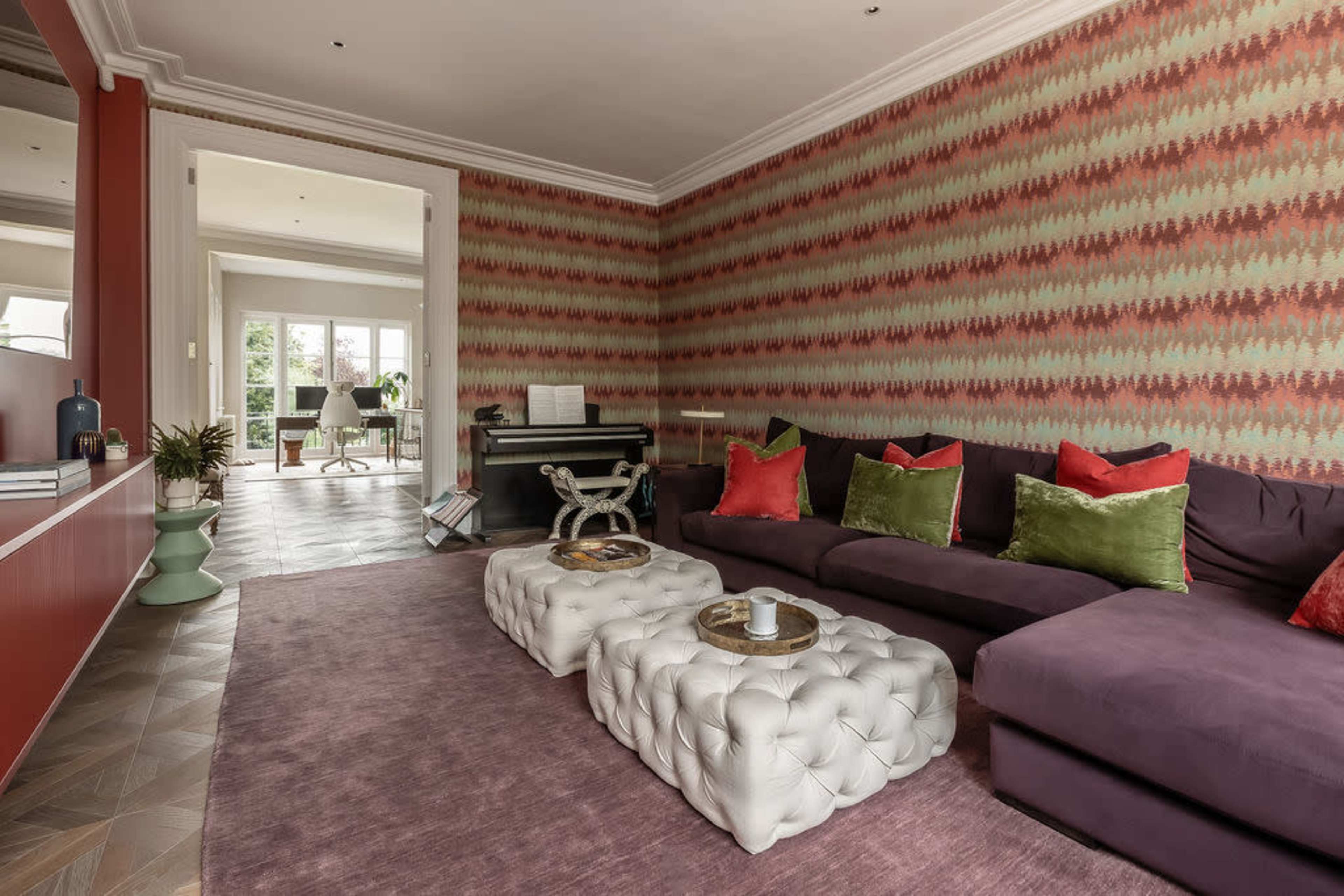 The living room features a purple sectional sofa, two tufted ottomans, and patterned wallpaper, with a view into an adjoining room.