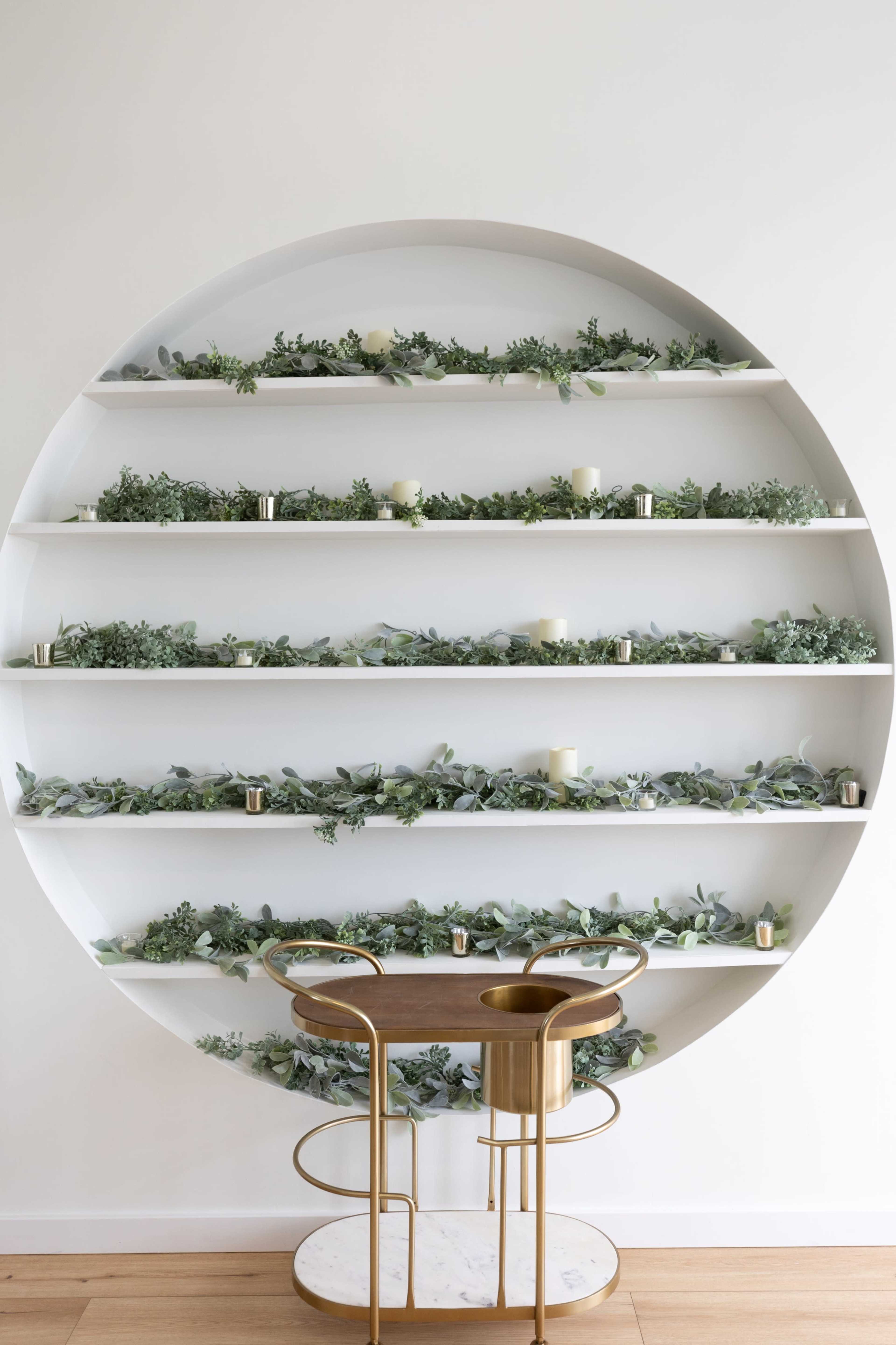 A circular white shelving unit displays several rows of greenery and candles, with a small round table and two chairs in front.